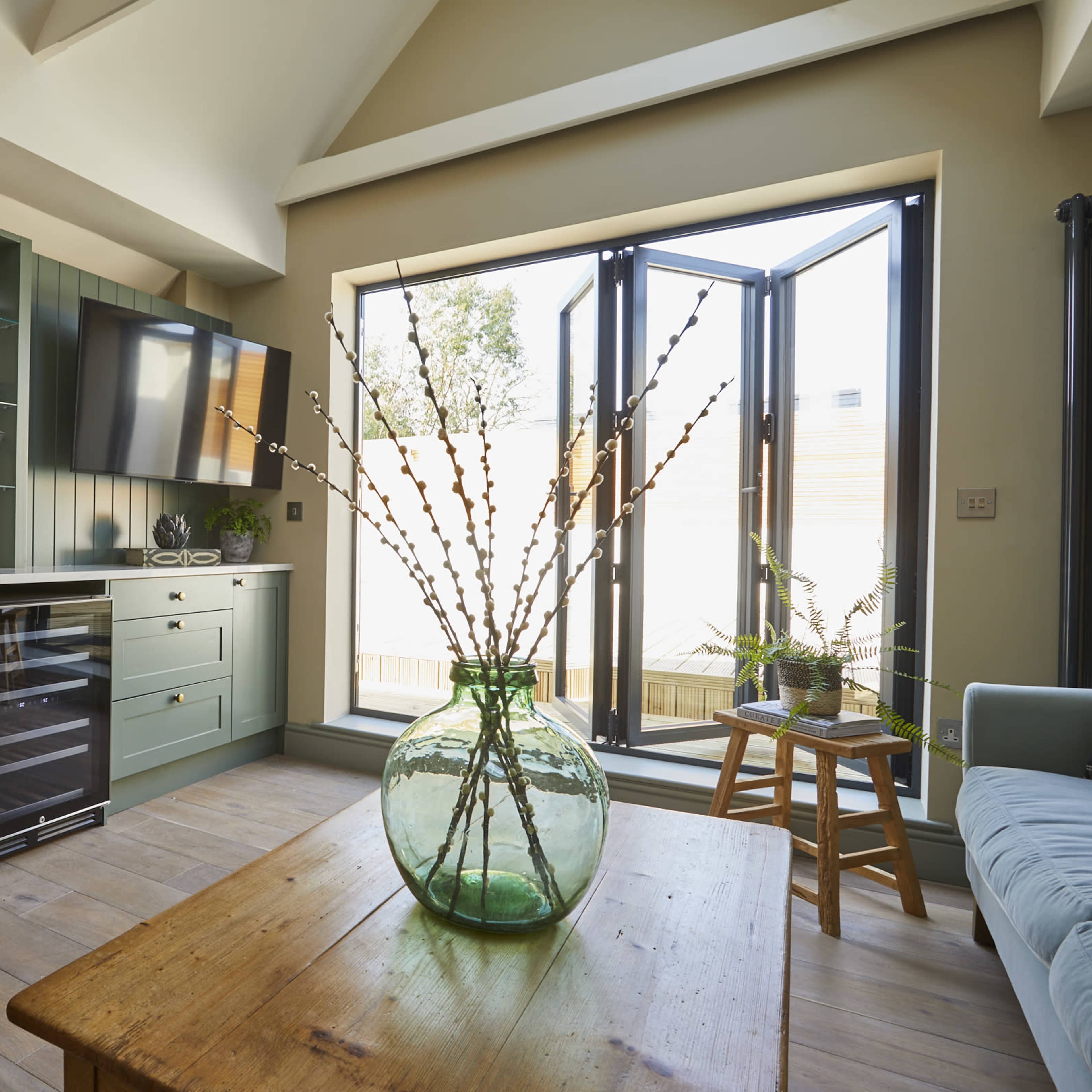A modern kitchen features a green cabinetry setup with a wine cooler, a glassware display, and a large window that opens to an outdoor area.