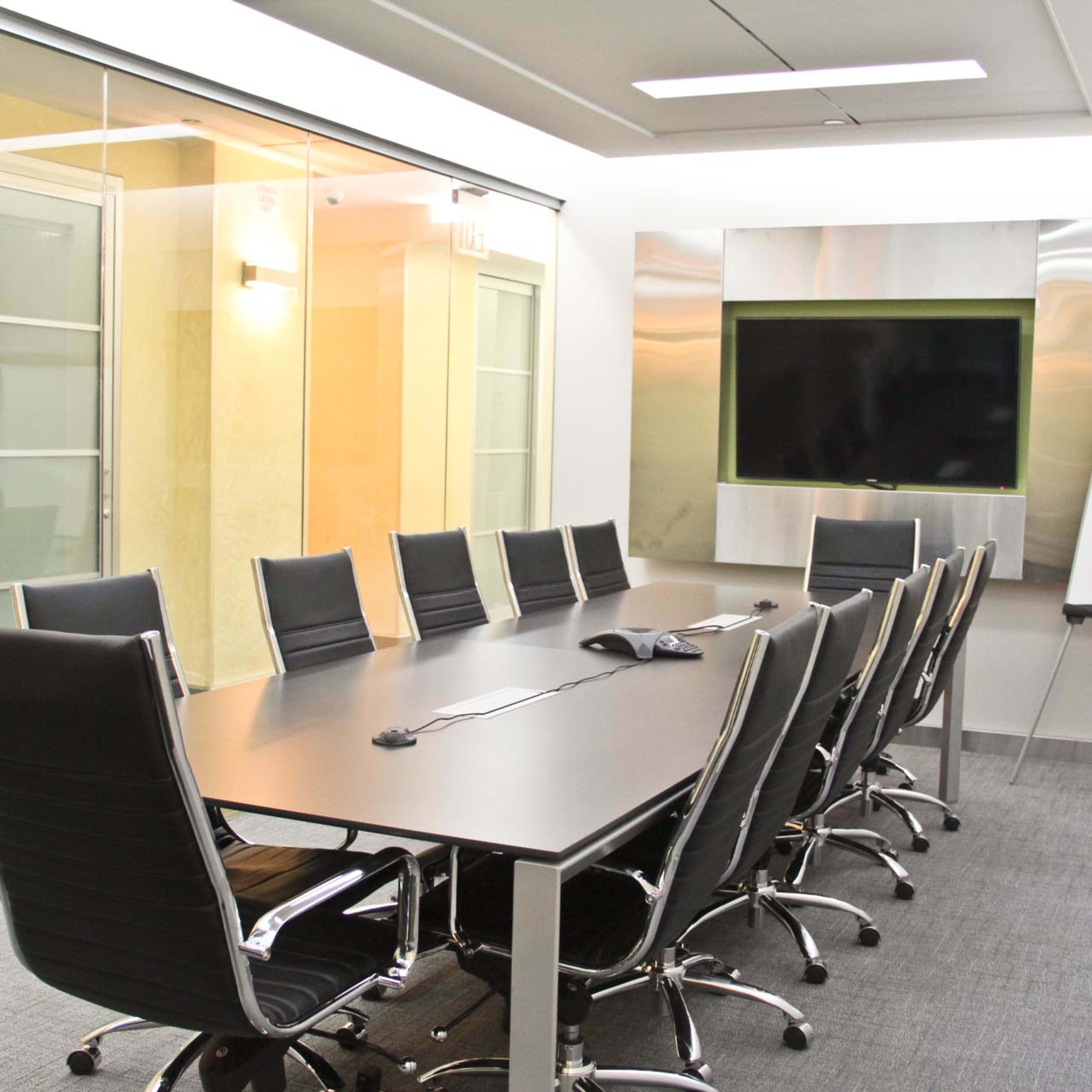 A conference room features a long table surrounded by black chairs, with a glass wall and a large screen at one end.
