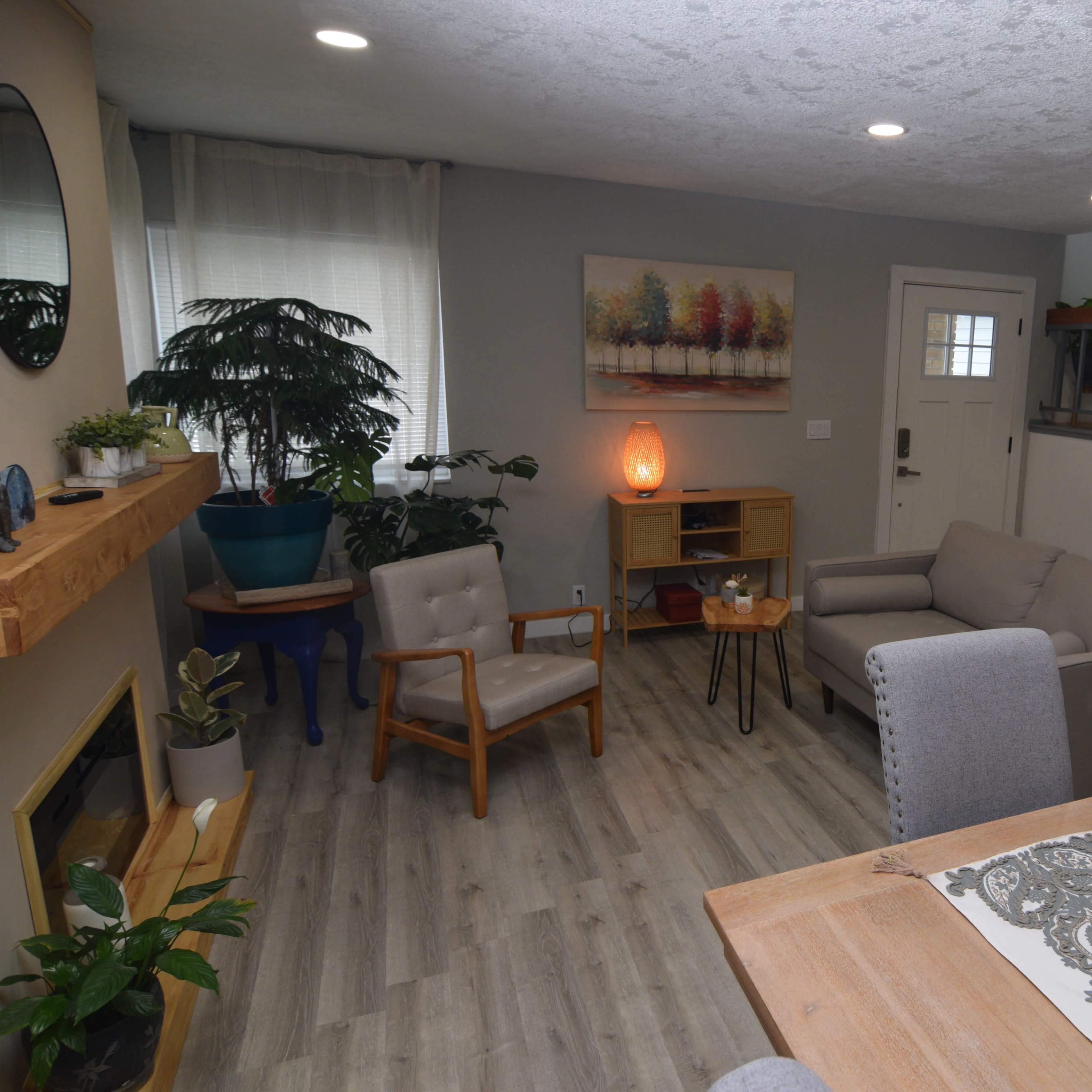 A modern living room with a comfortable seating arrangement, a wooden dining table, a small bookshelf, and a staircase in the background.