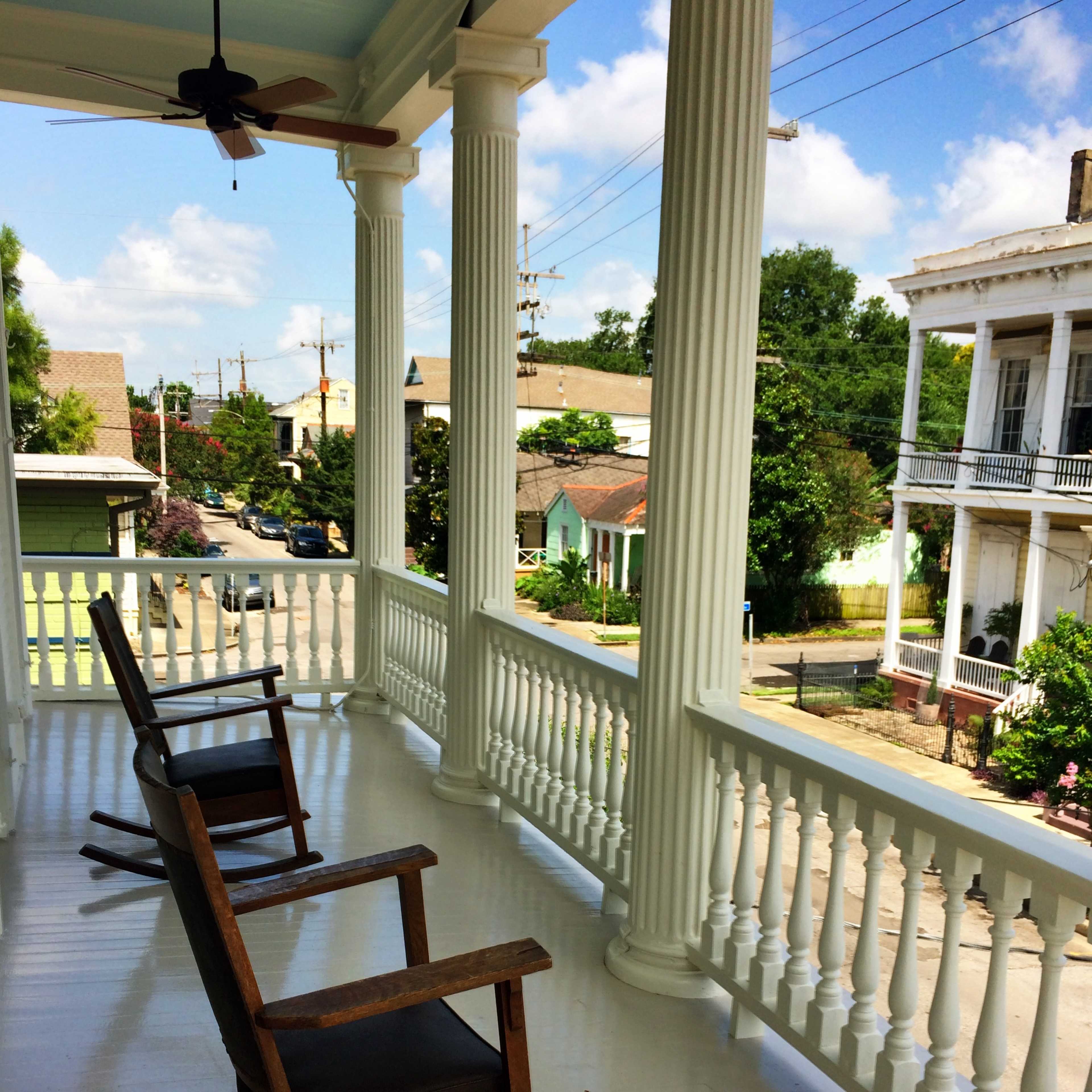 A porch with two rocking chairs overlooking a street lined with houses and trees under a blue sky.