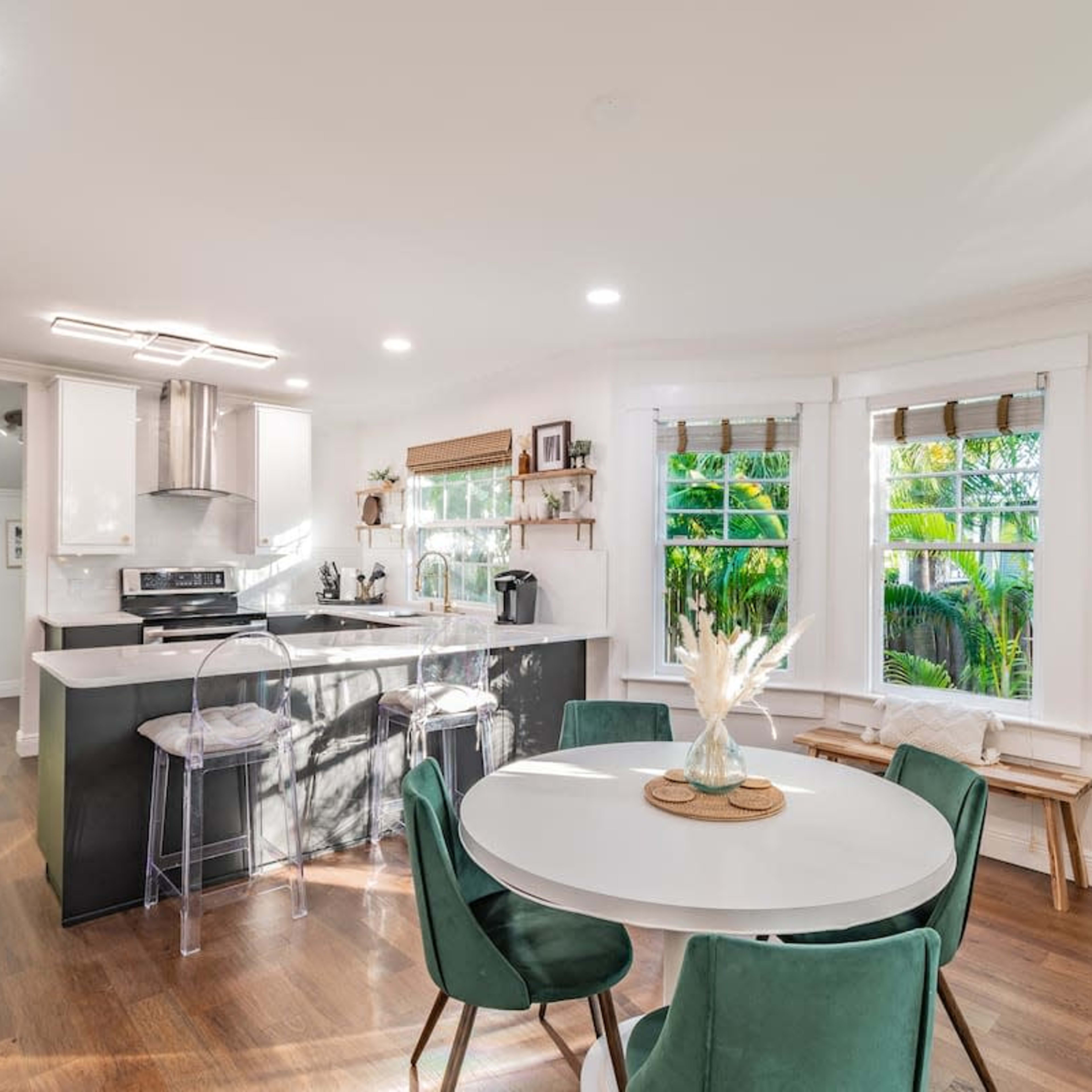 A bright, modern kitchen and dining area with a round table, green chairs, and large windows overlooking a garden.
