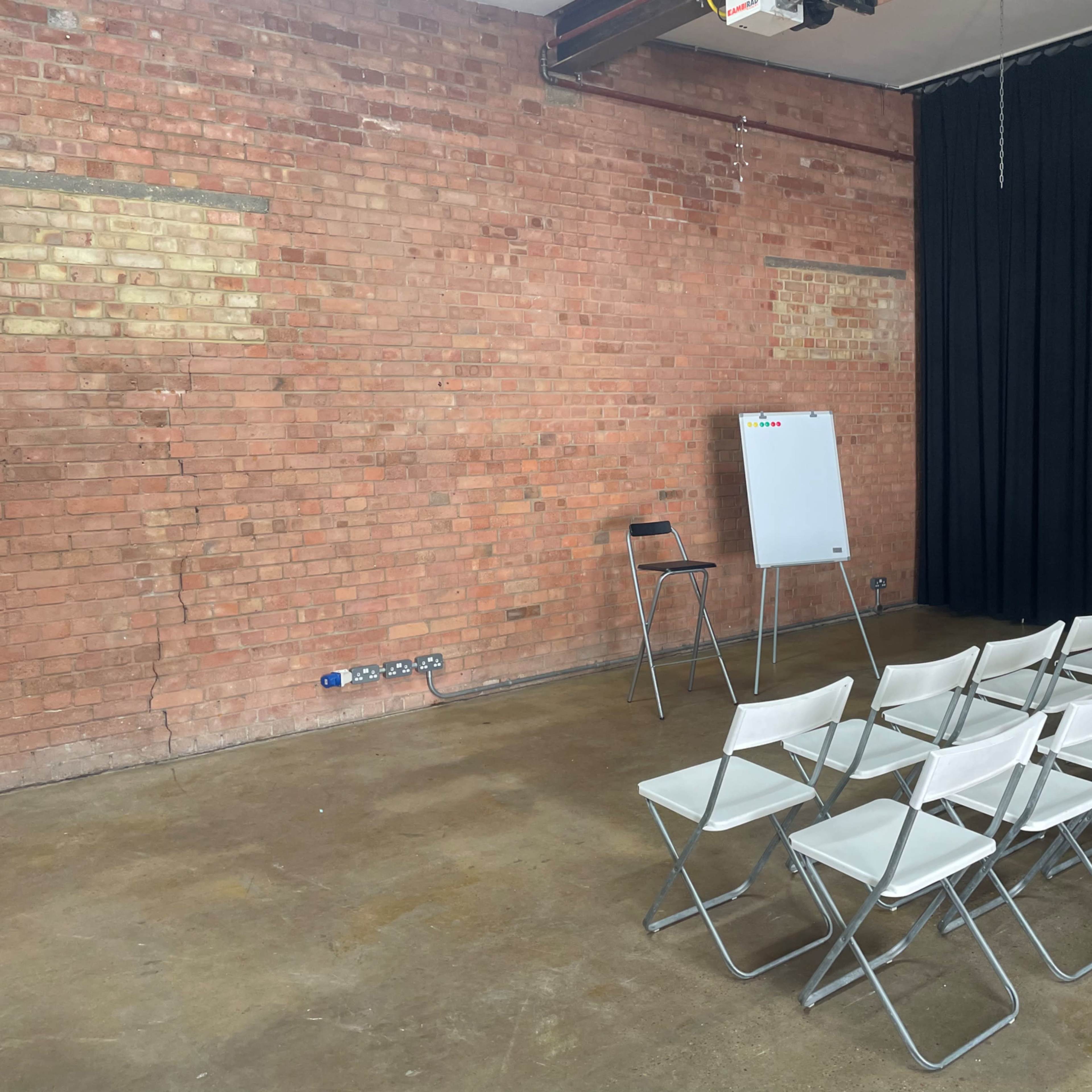 A room with exposed brick walls features a row of white folding chairs facing a blank flip chart on a stand beside a single black chair.