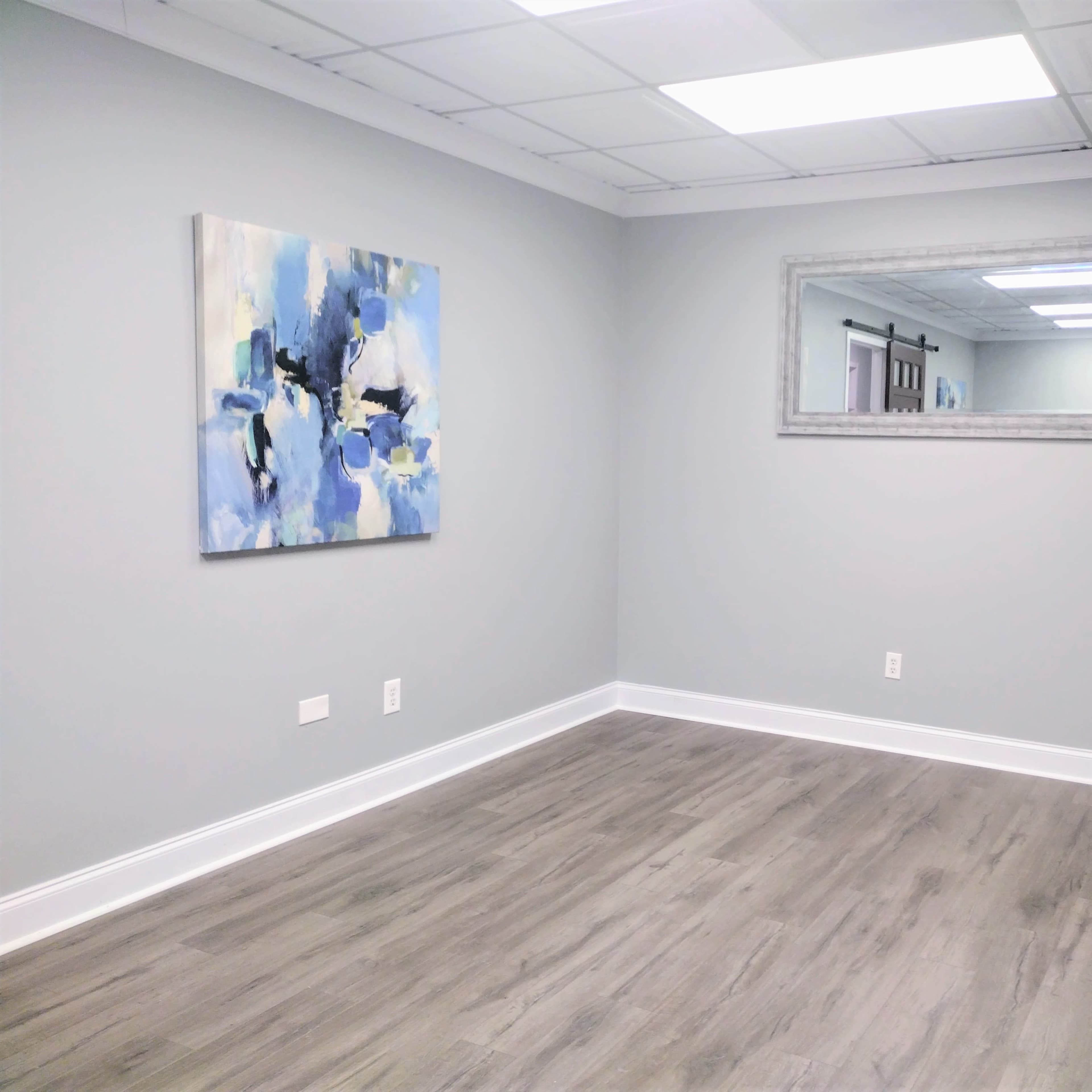 A light gray room features a large abstract painting on one wall, a rectangular mirror on the opposite wall, and two decorative baskets in the corner.