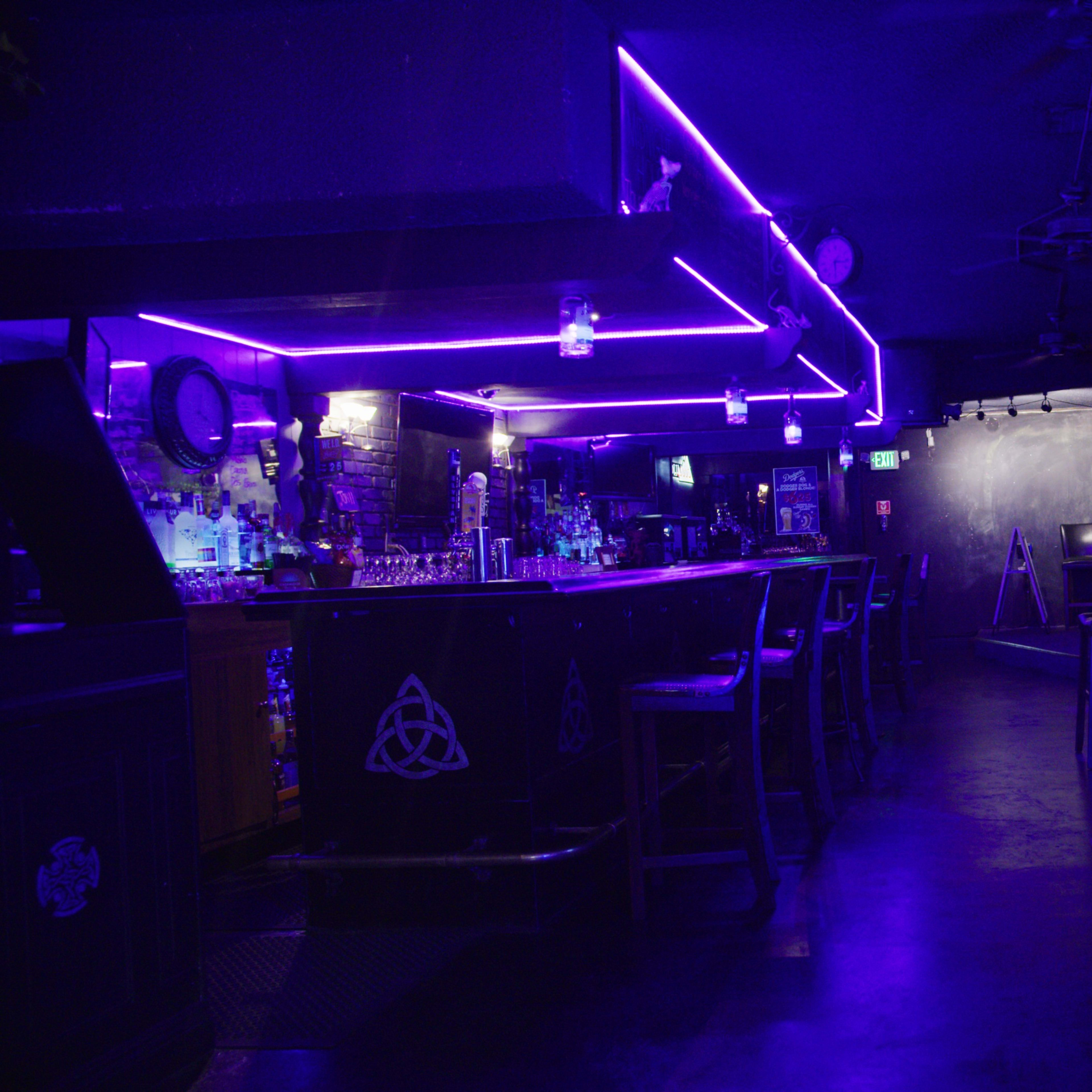 A dimly lit bar features a counter with glowing purple neon lights and several empty stools.