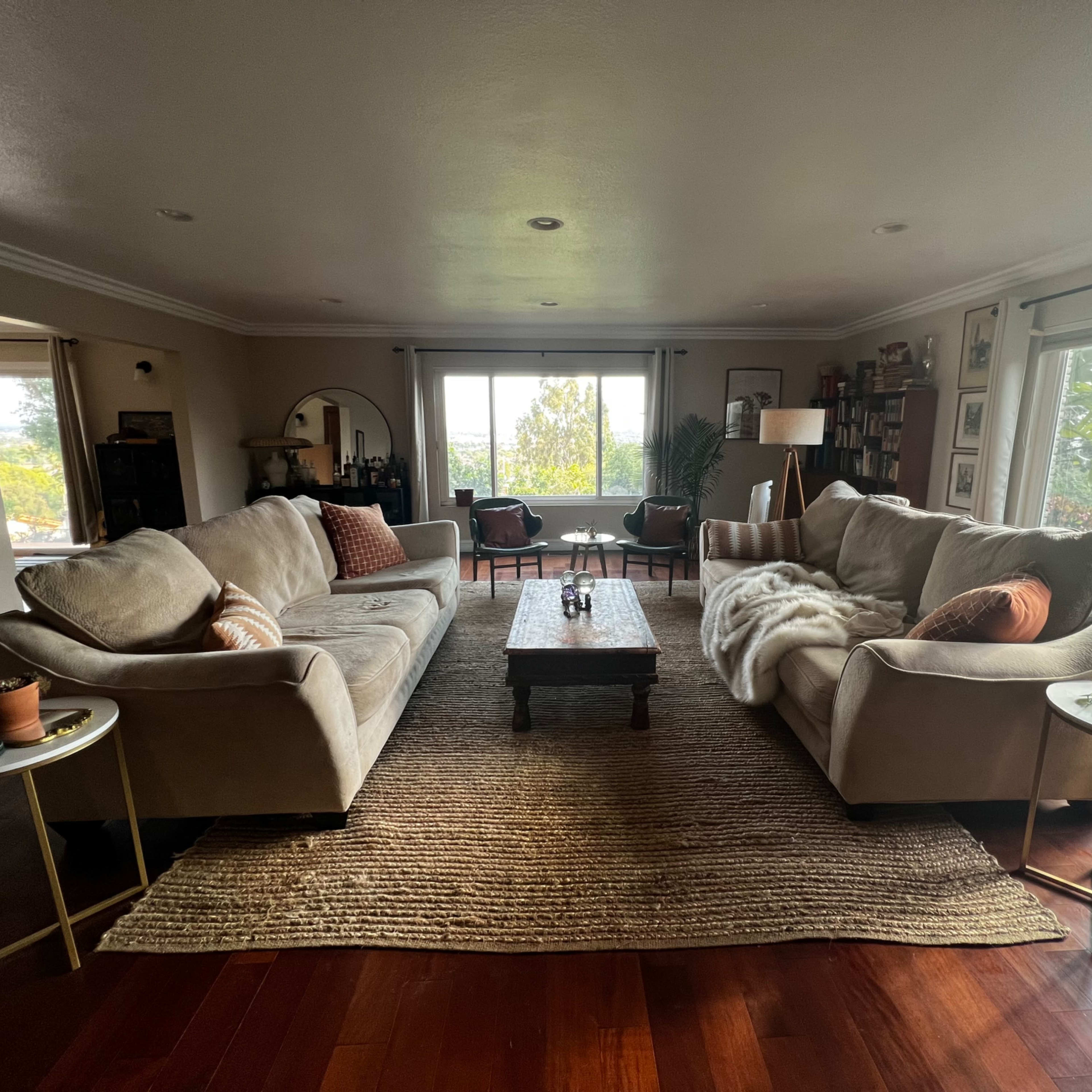 A spacious living room features two beige sofas facing a wooden coffee table, with large windows showcasing outdoor greenery.