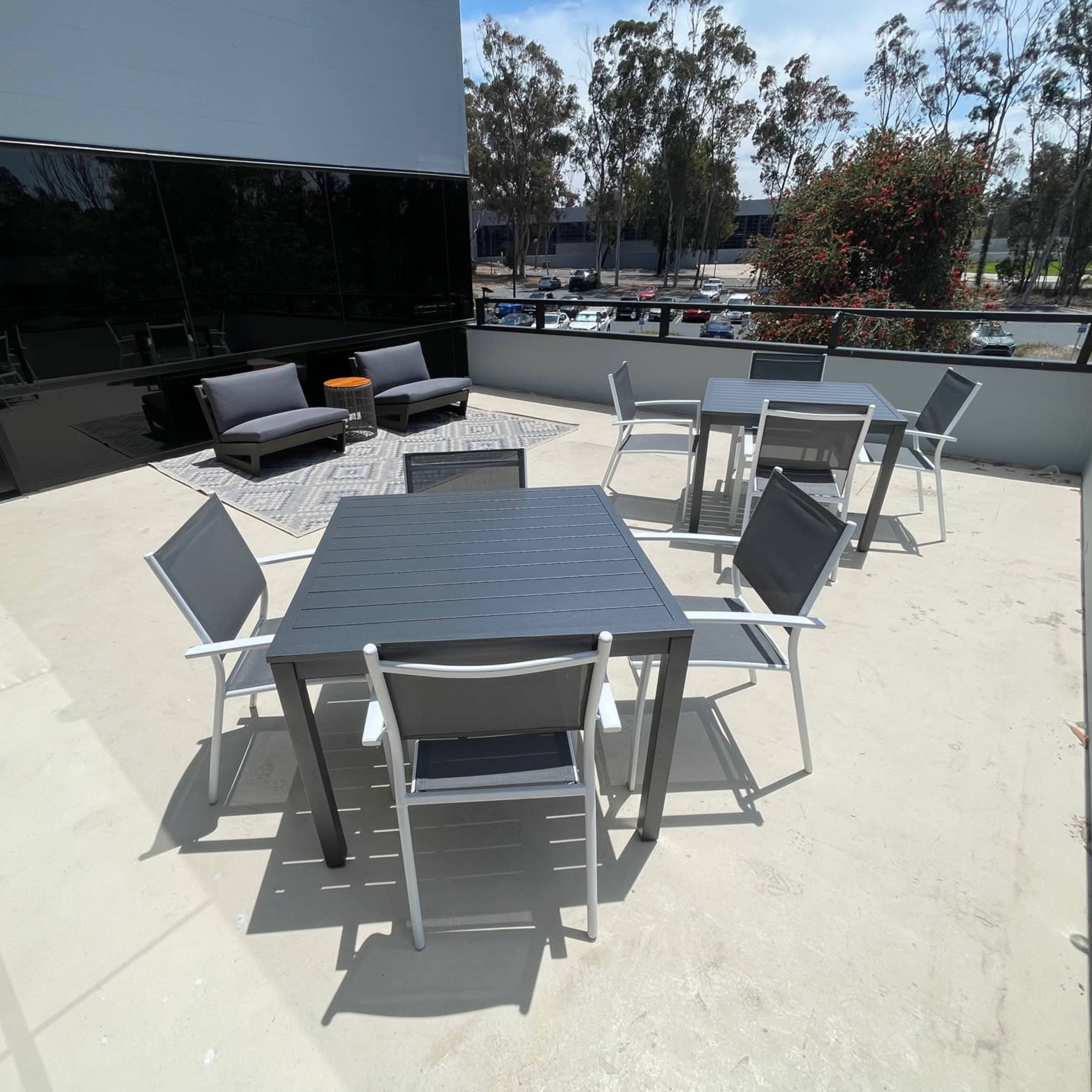 A spacious outdoor terrace with several tables and chairs, a rug, and seating areas surrounded by trees and buildings.