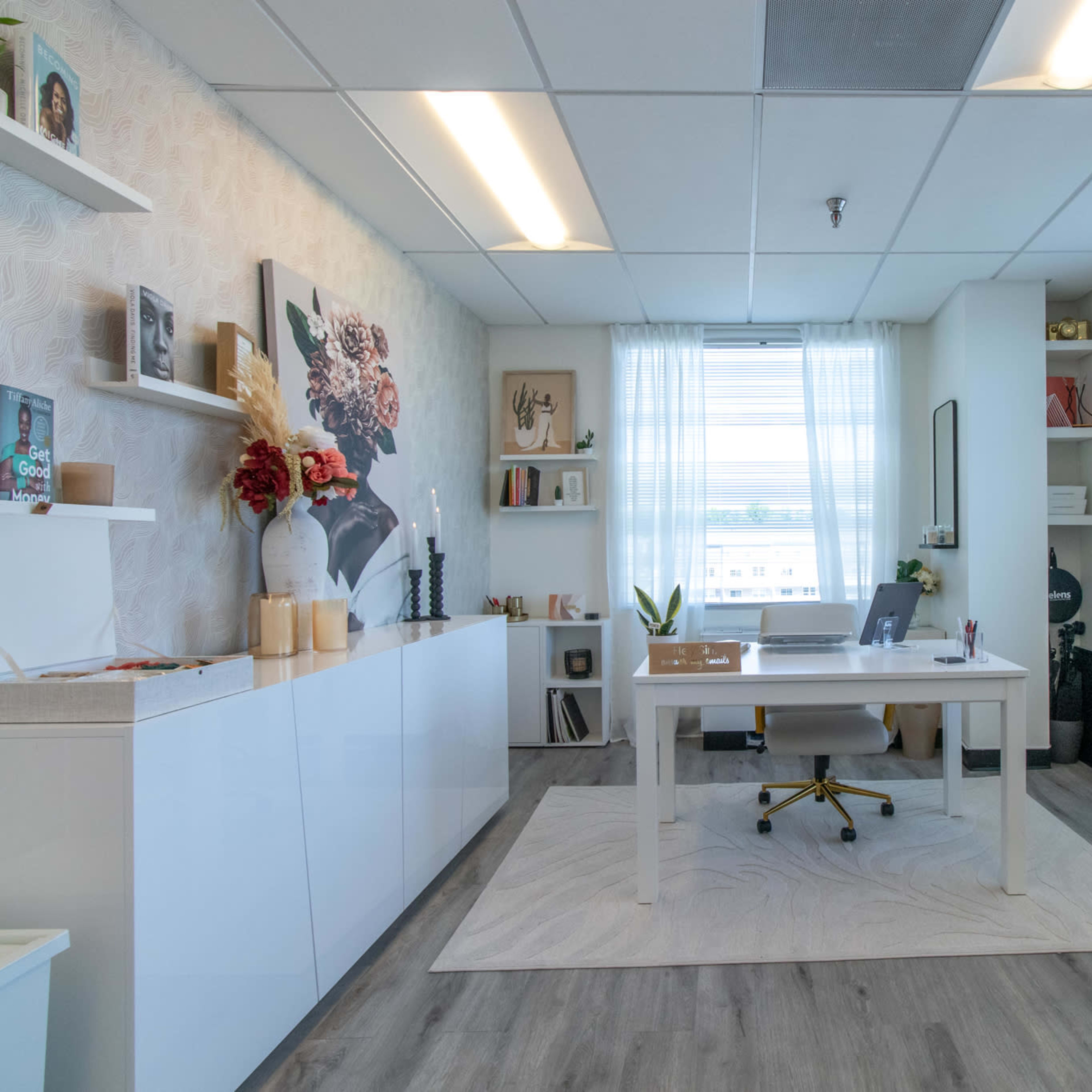 A modern office space features a white desk with a chair, surrounded by shelves displaying decor and books, and large windows allowing natural light to enter.