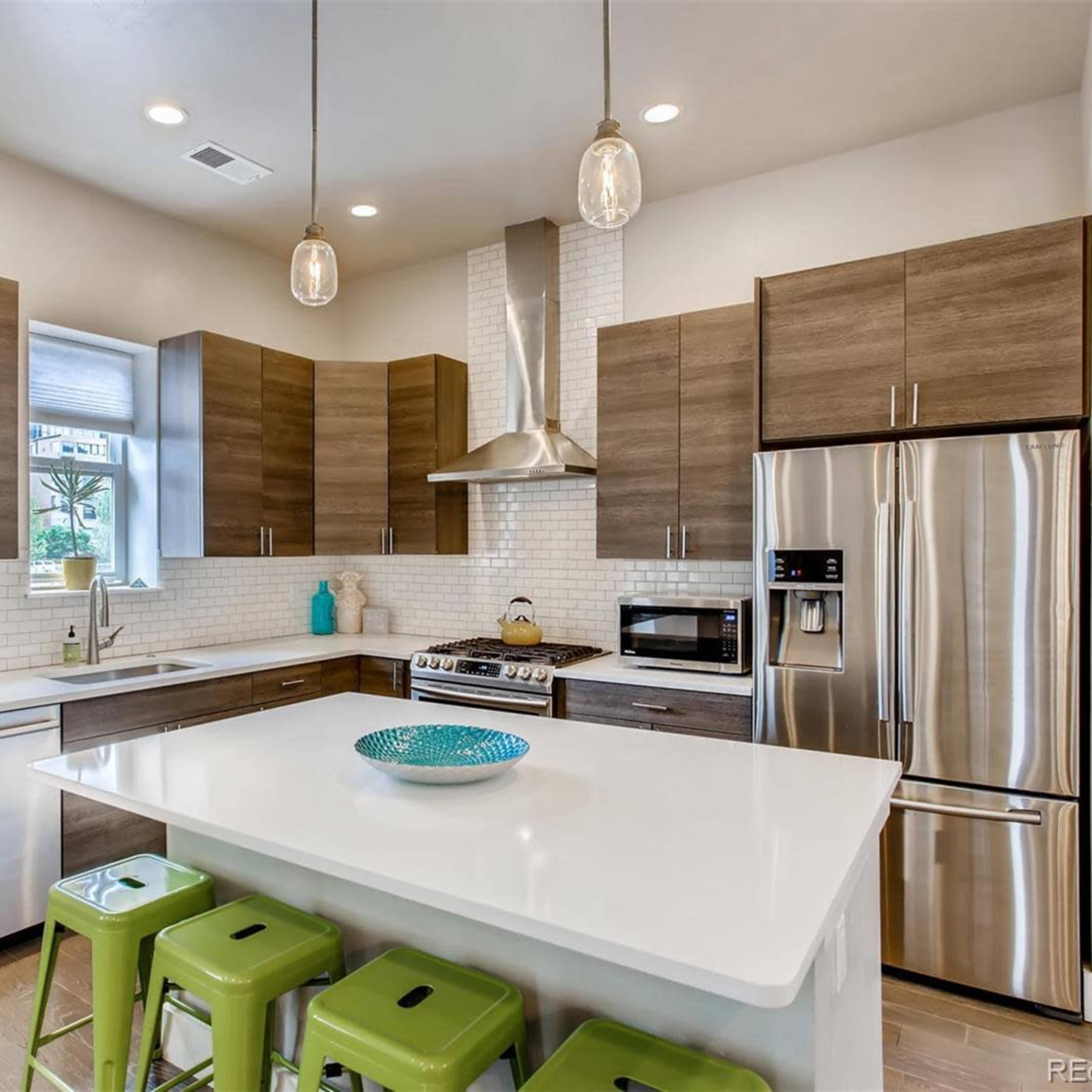A modern kitchen featuring brown wood cabinets, white subway tile backsplash, stainless steel appliances, and a large island with green stools.