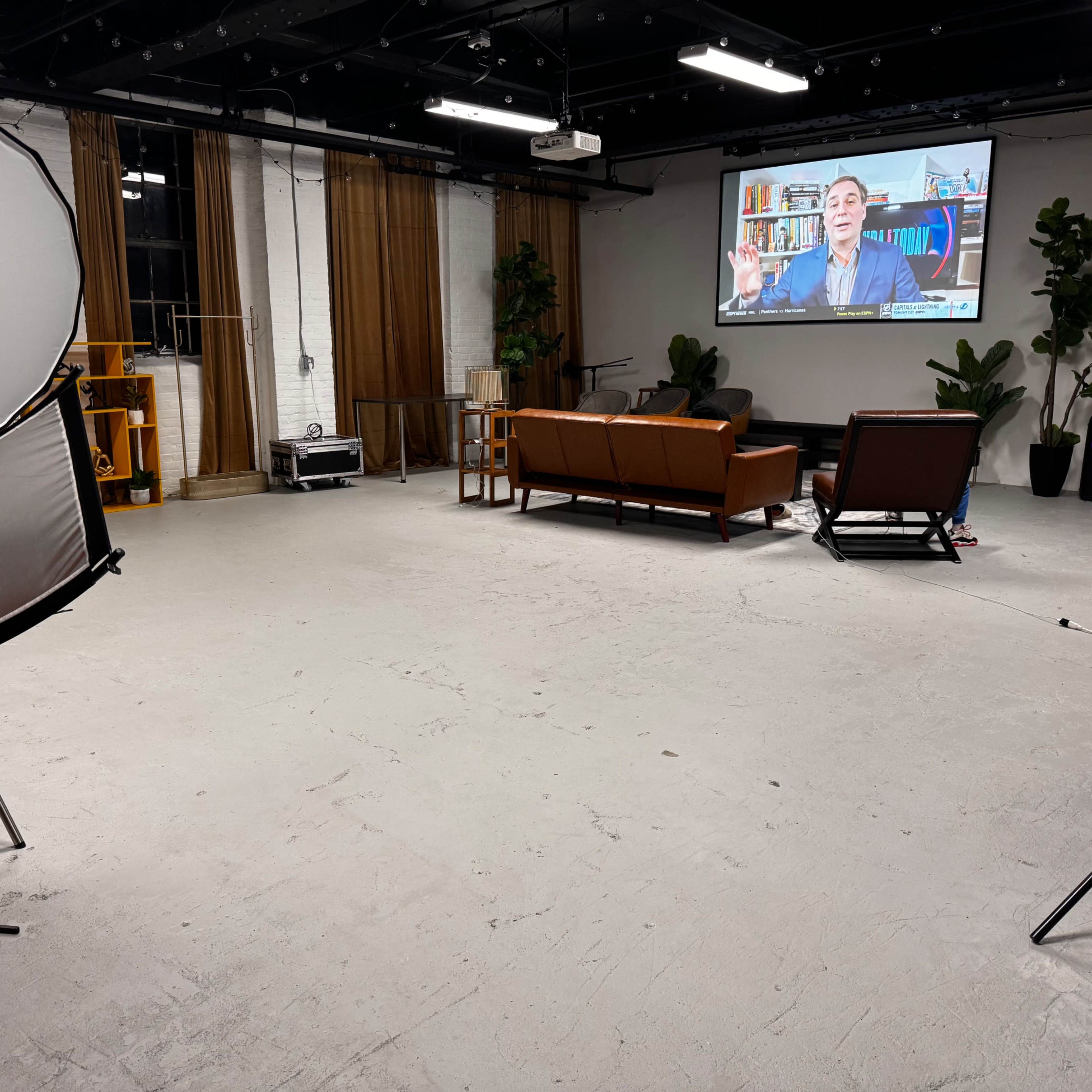 A spacious, well-lit studio with a large screen displaying a man speaking, two brown couches, and various plants arranged around the room.