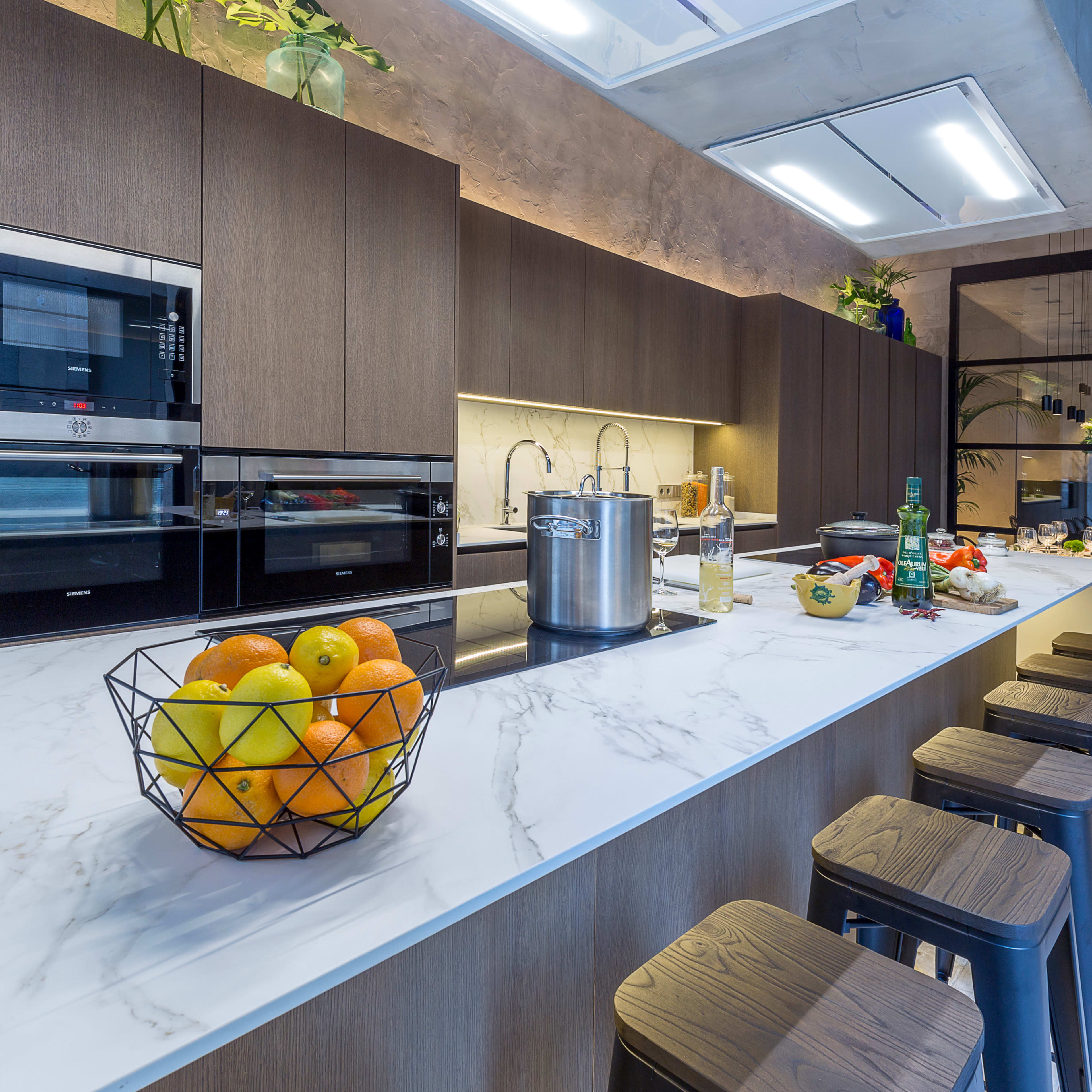 A modern kitchen features a marble island with a fruit bowl, sleek wooden cabinetry, and stainless steel appliances.