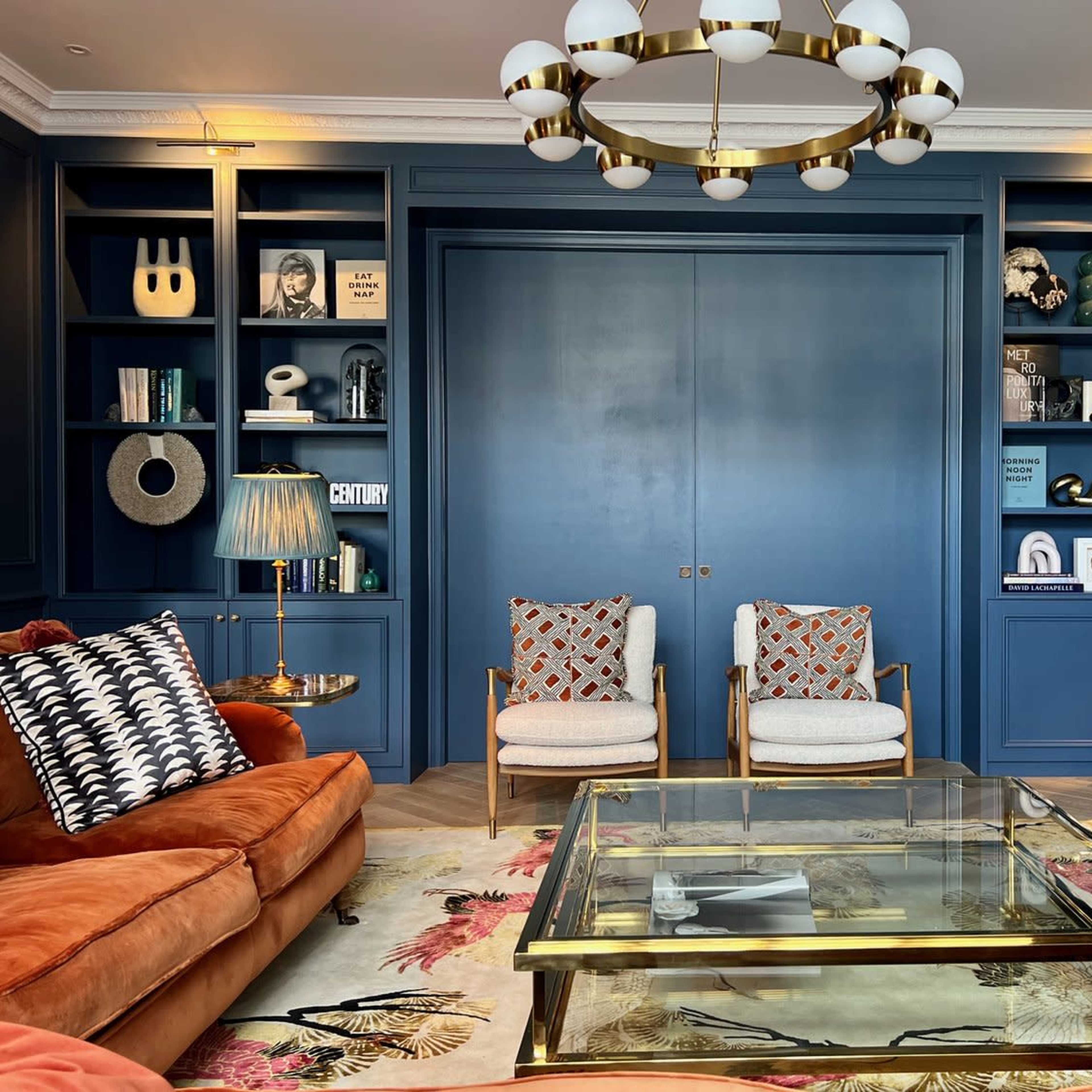 A stylish living room with a navy blue feature wall, a glass coffee table, two armchairs, a large orange sofa, and shelves filled with decorative items and books.