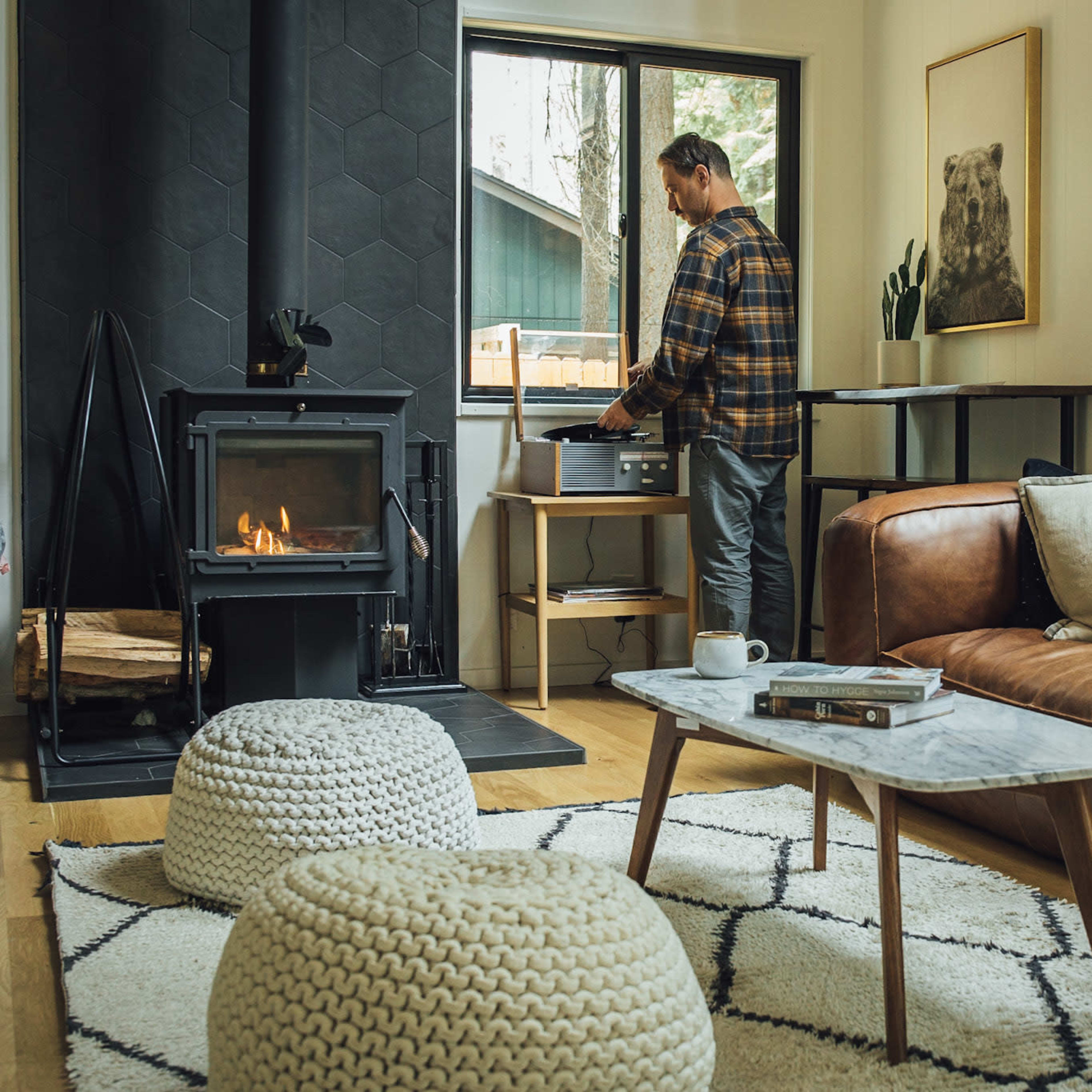 A man stands by a record player next to a large window in a cozy living room with a fireplace and a leather sofa.