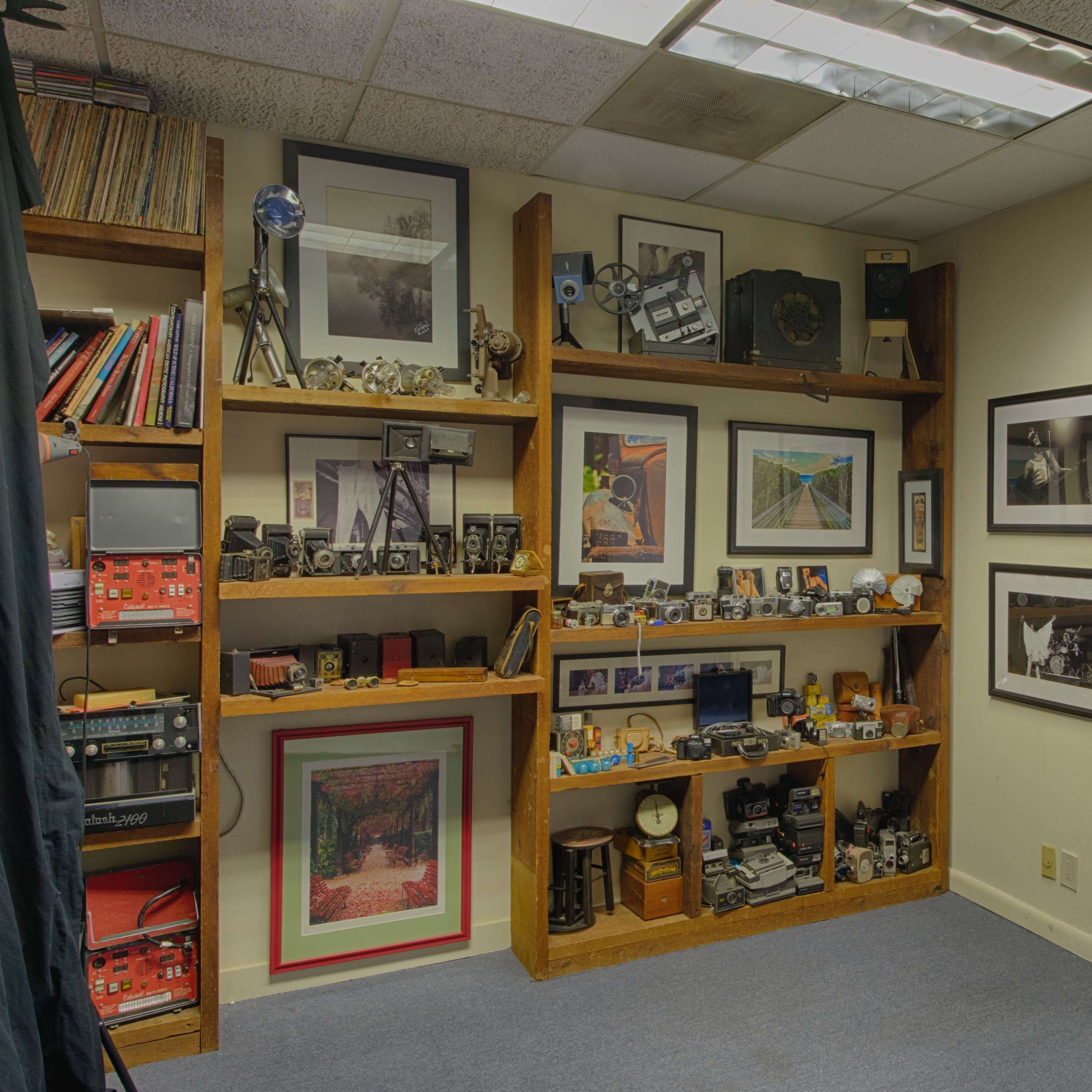 A corner of an office with wooden shelves displaying various photographs, cameras, and decorative items, adjacent to an open doorway leading to another room.