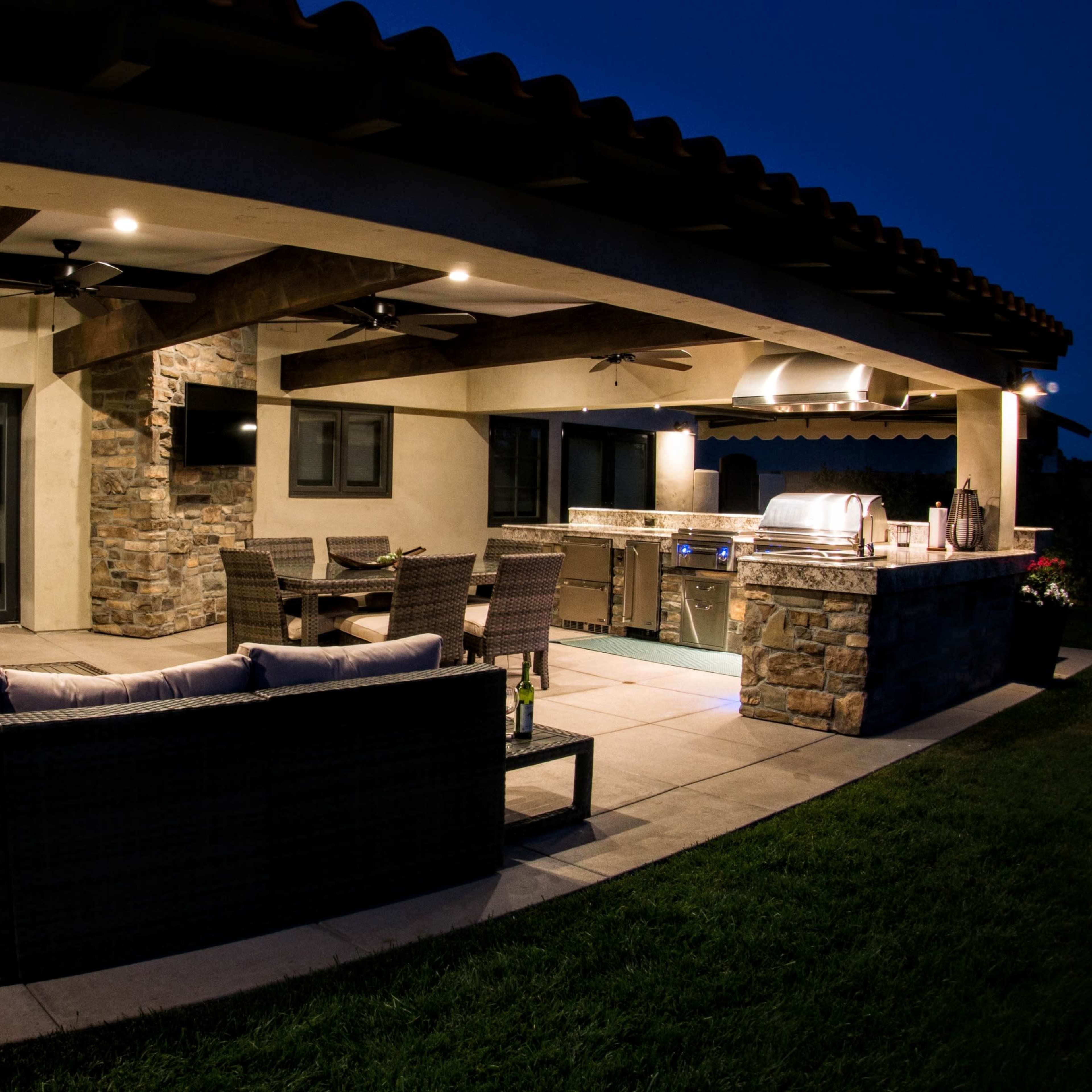 An outdoor living space featuring a covered patio with a seating area, a stone grill station, and large glass doors leading to an interior space.