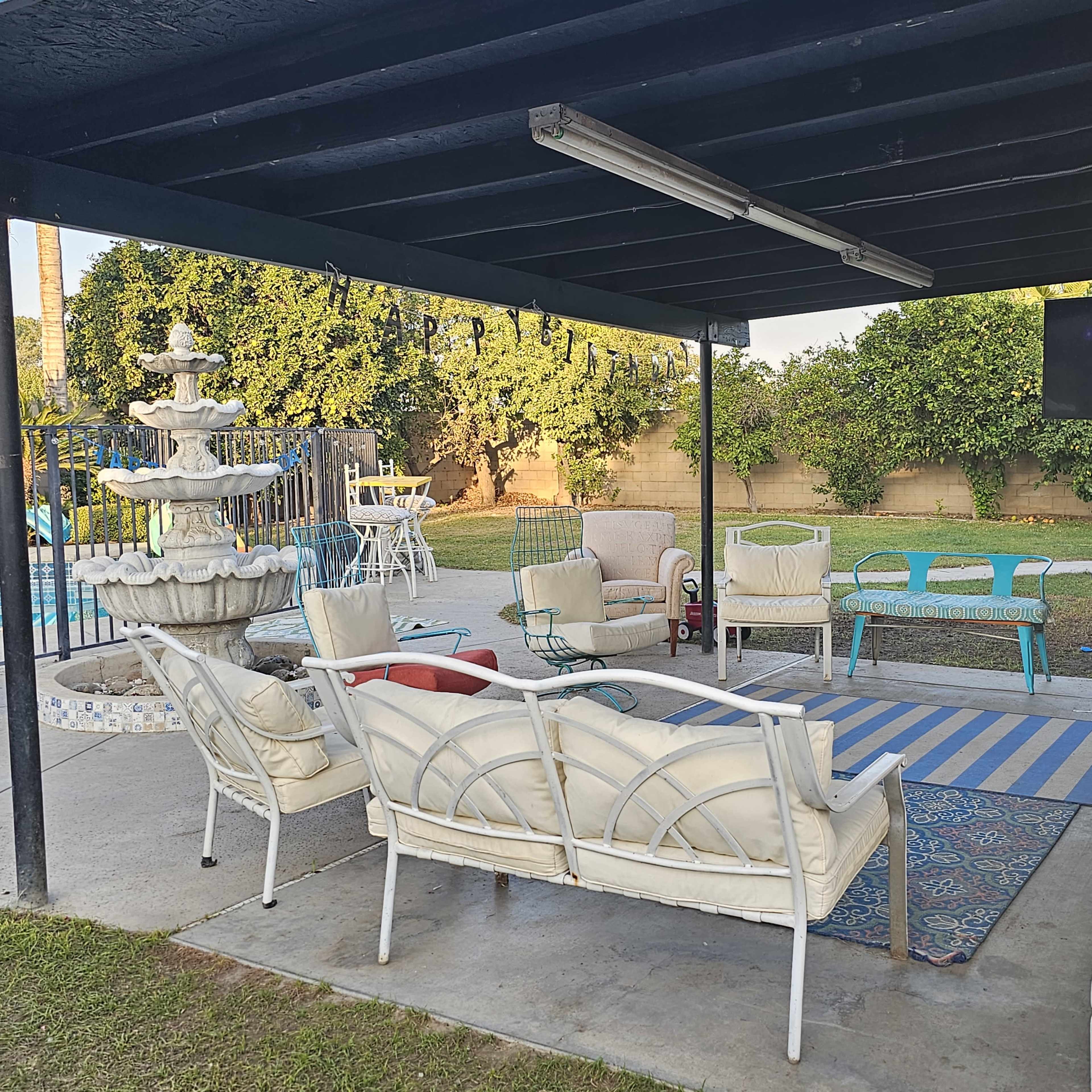 A covered outdoor seating area with white patio furniture, a fountain, a swimming pool in the background, and a television mounted on the wall.