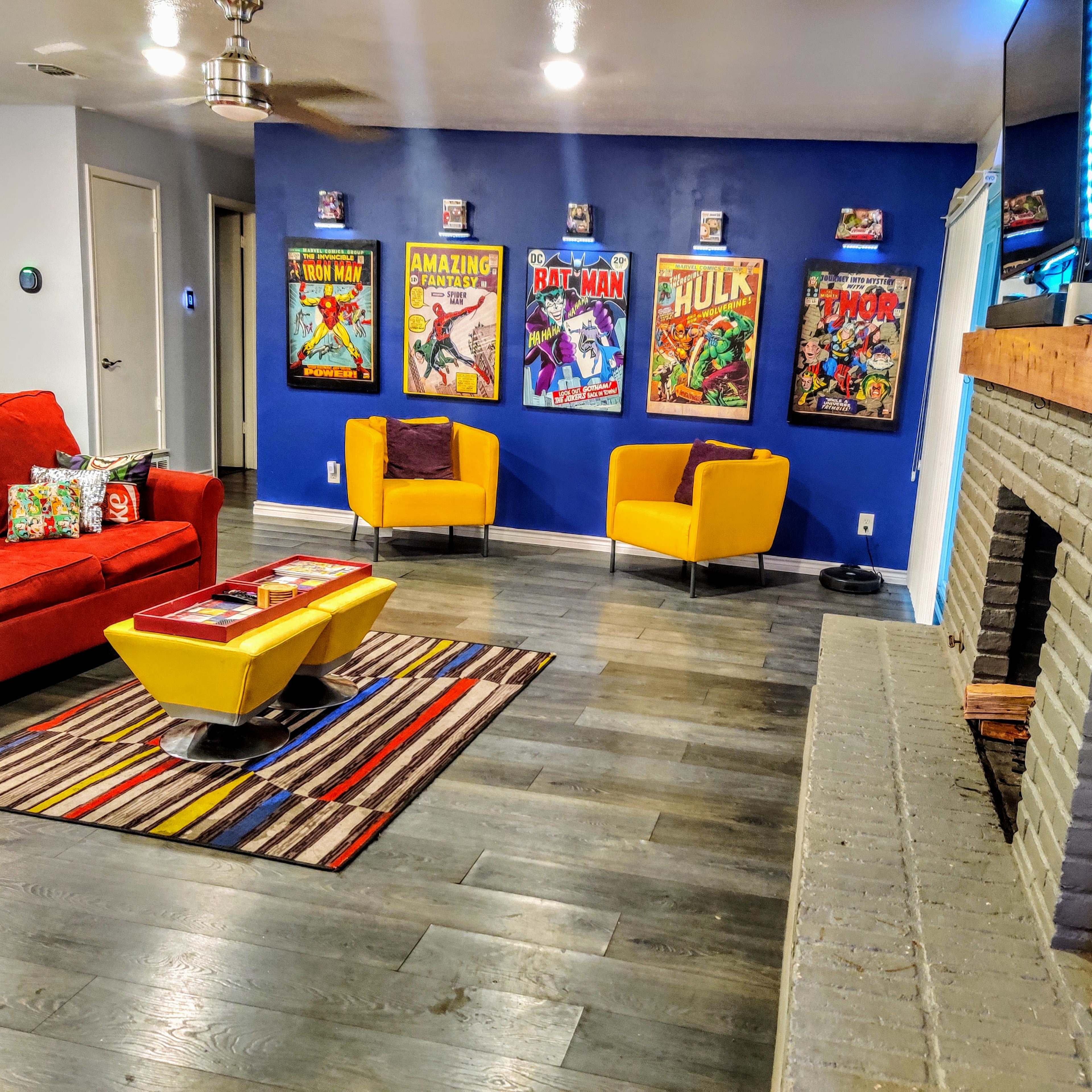 The living room features a red sofa, yellow chairs, a patterned rug, and a wall adorned with framed comic book posters.