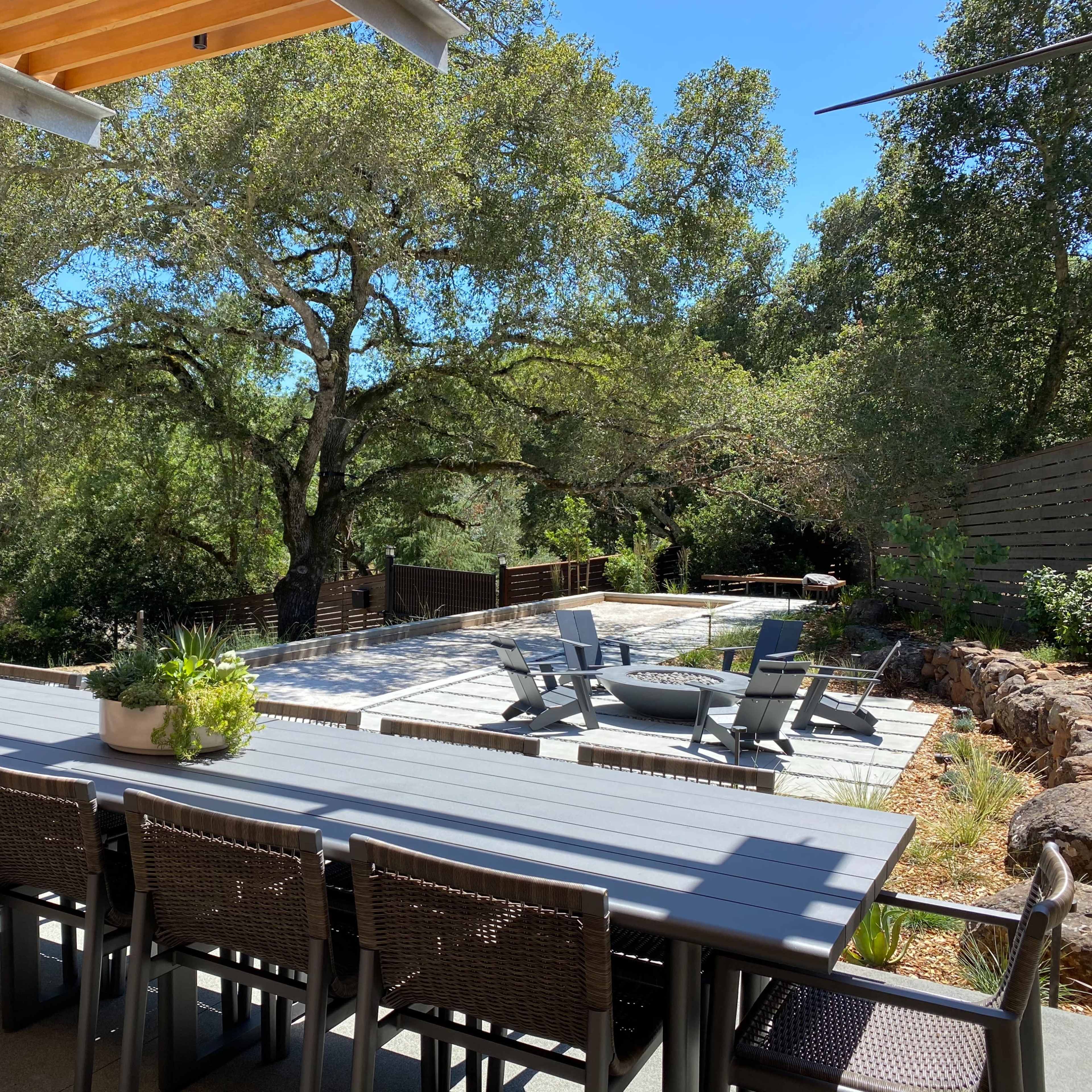 A long dining table with chairs is set on a patio overlooking a swimming pool surrounded by greenery and trees.