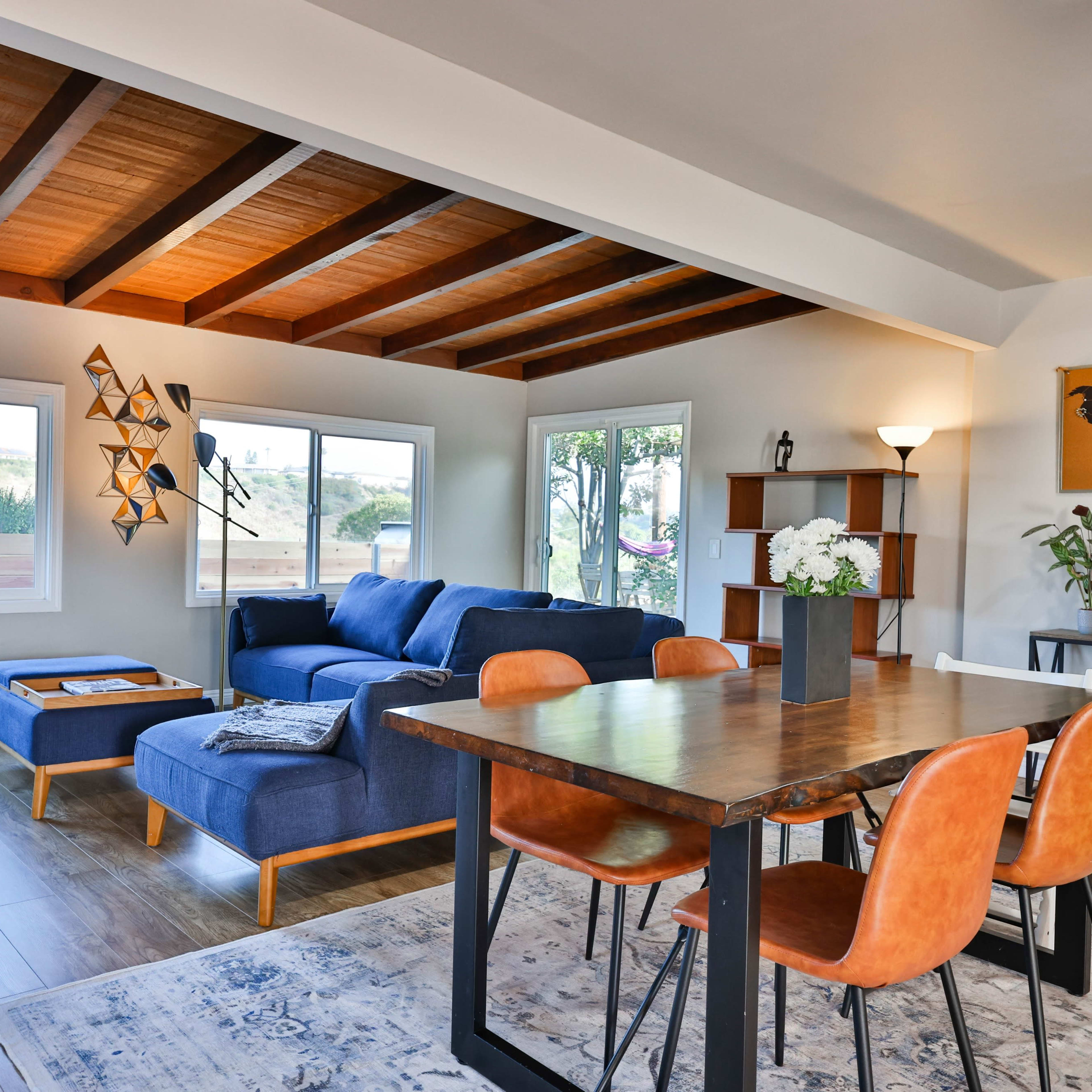 A modern living and dining area featuring a blue sofa, a wooden dining table with orange chairs, and large windows that allow natural light to fill the space.