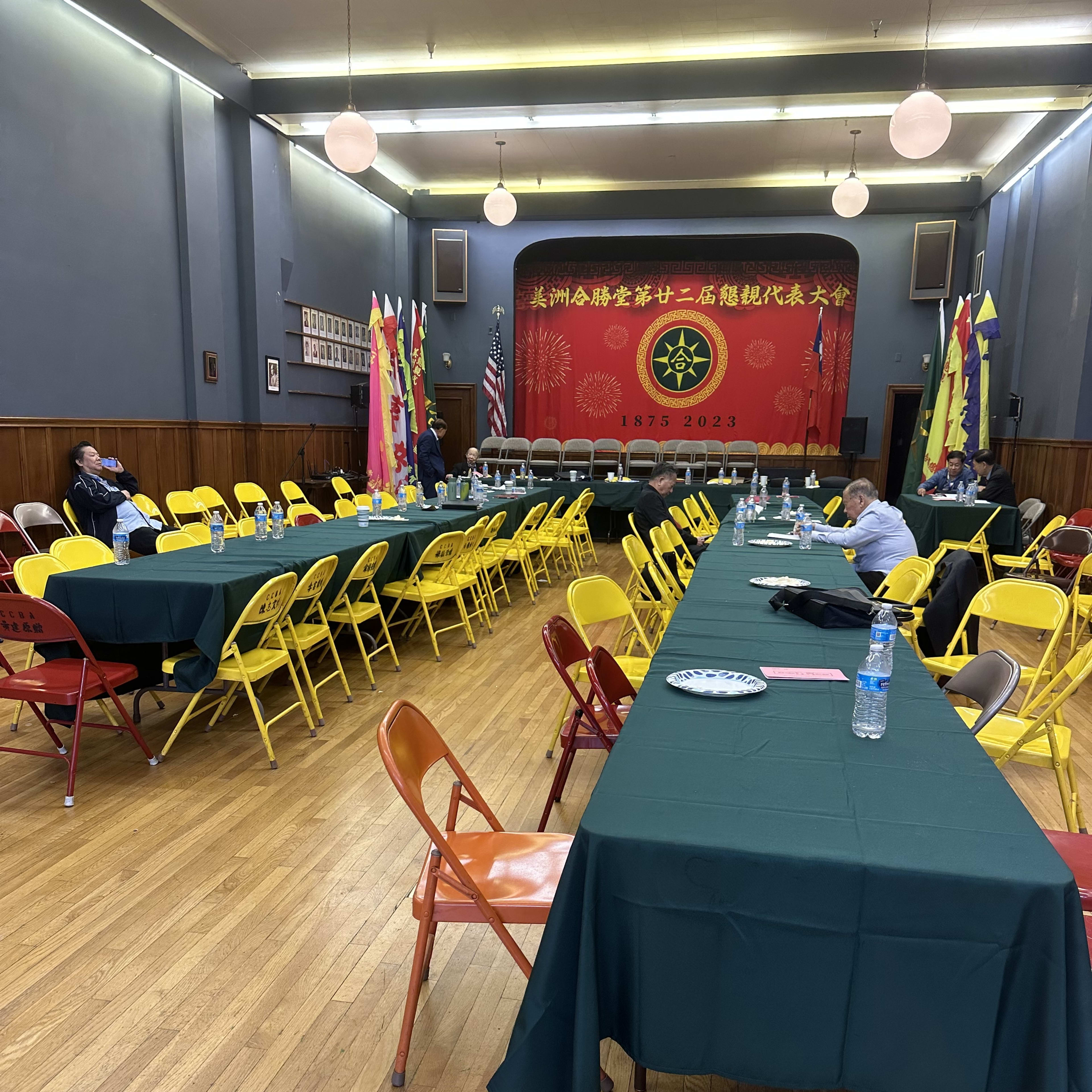 A large hall with yellow and green folding chairs arranged around several long tables, featuring a red backdrop with decorative banners and flags.