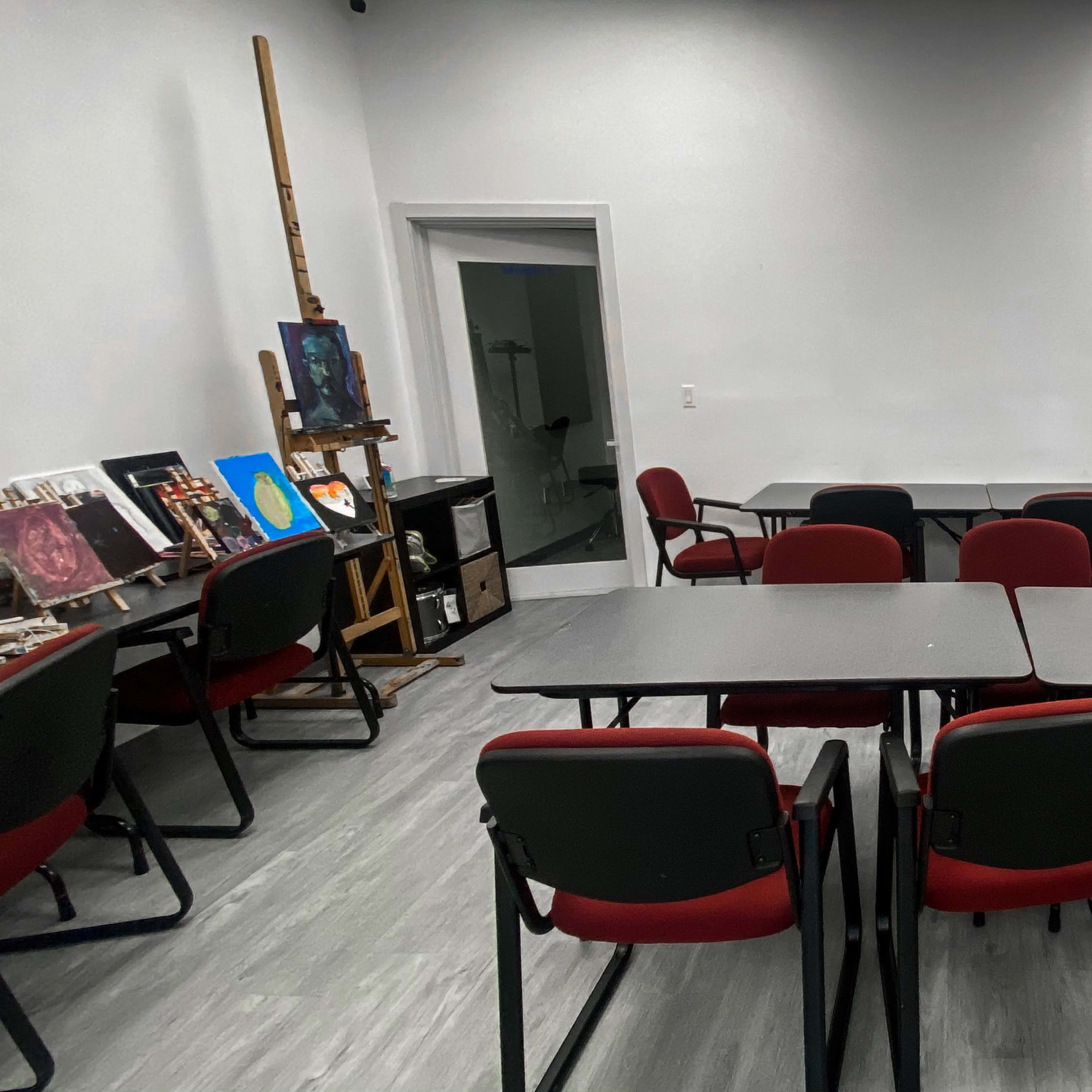 A small classroom with several tables and chairs arranged facing a blank wall, alongside an easel holding artwork.