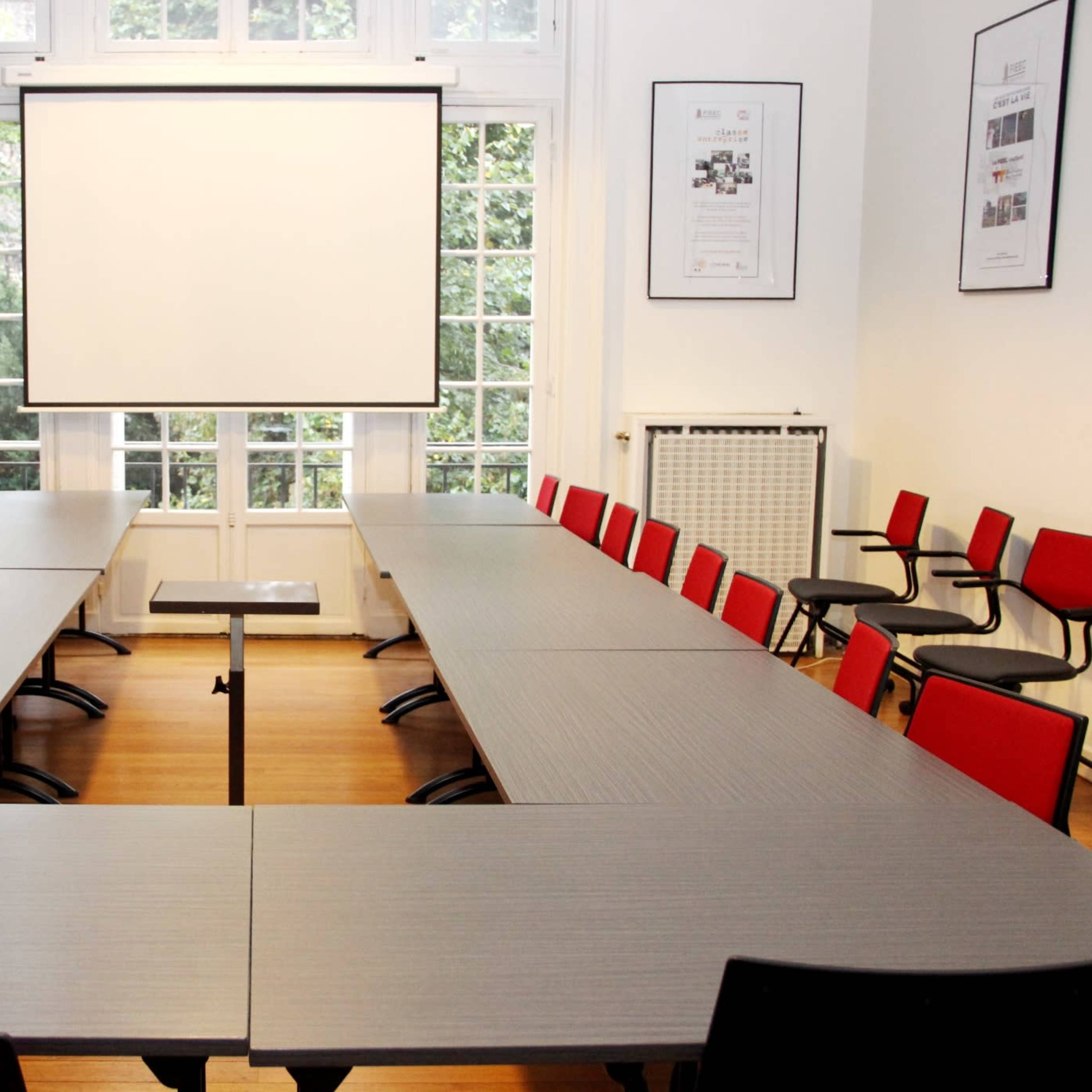 A large meeting room features two sets of rectangular tables arranged in a U-shape, surrounded by red chairs, with a blank projection screen at one end and large windows providing natural light.