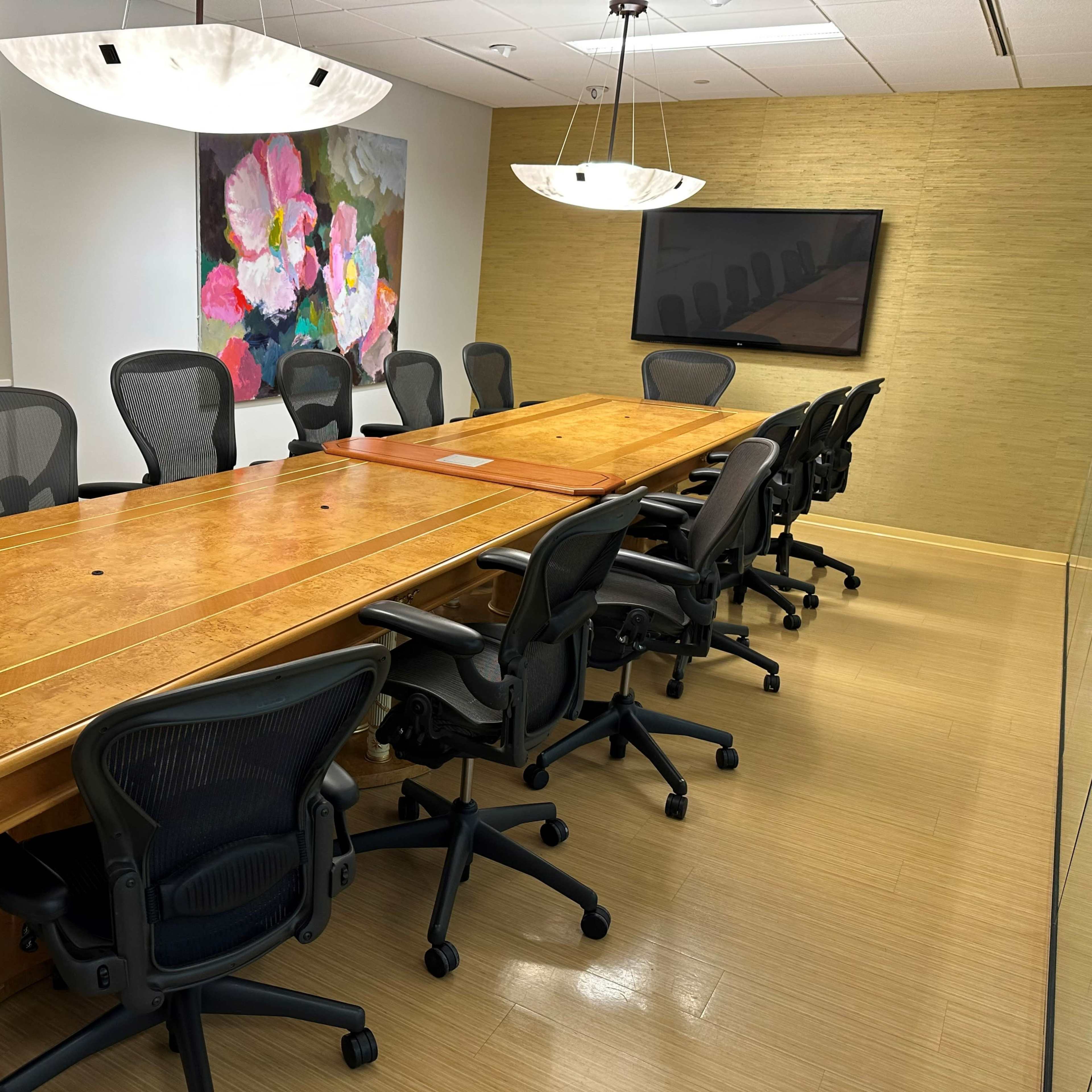 A modern conference room featuring a large wooden table surrounded by black office chairs, with artwork on the walls and a wall-mounted screen.