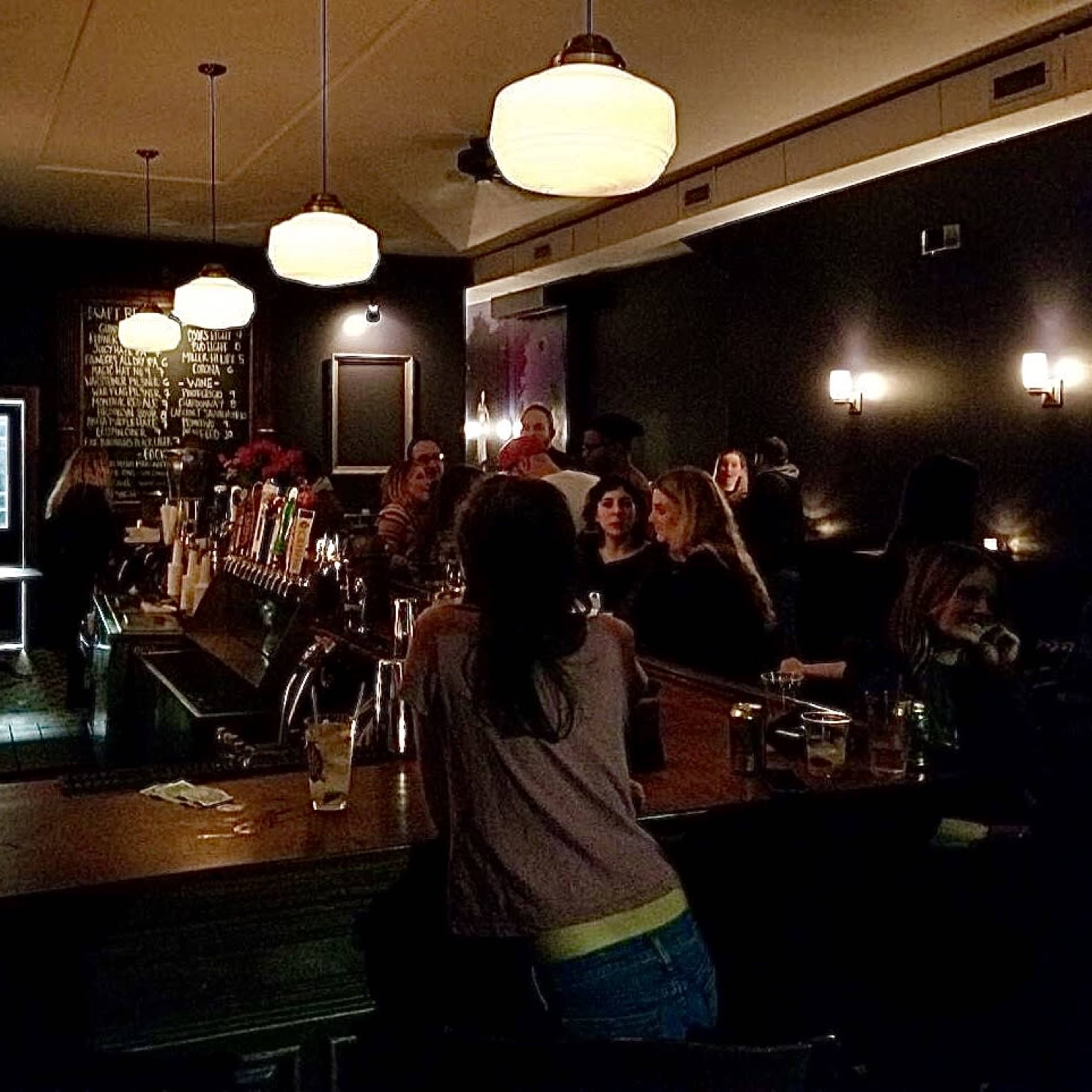 A dimly lit bar features a wooden counter with patrons seated, while flowers decorate the space and the walls display a drink menu.