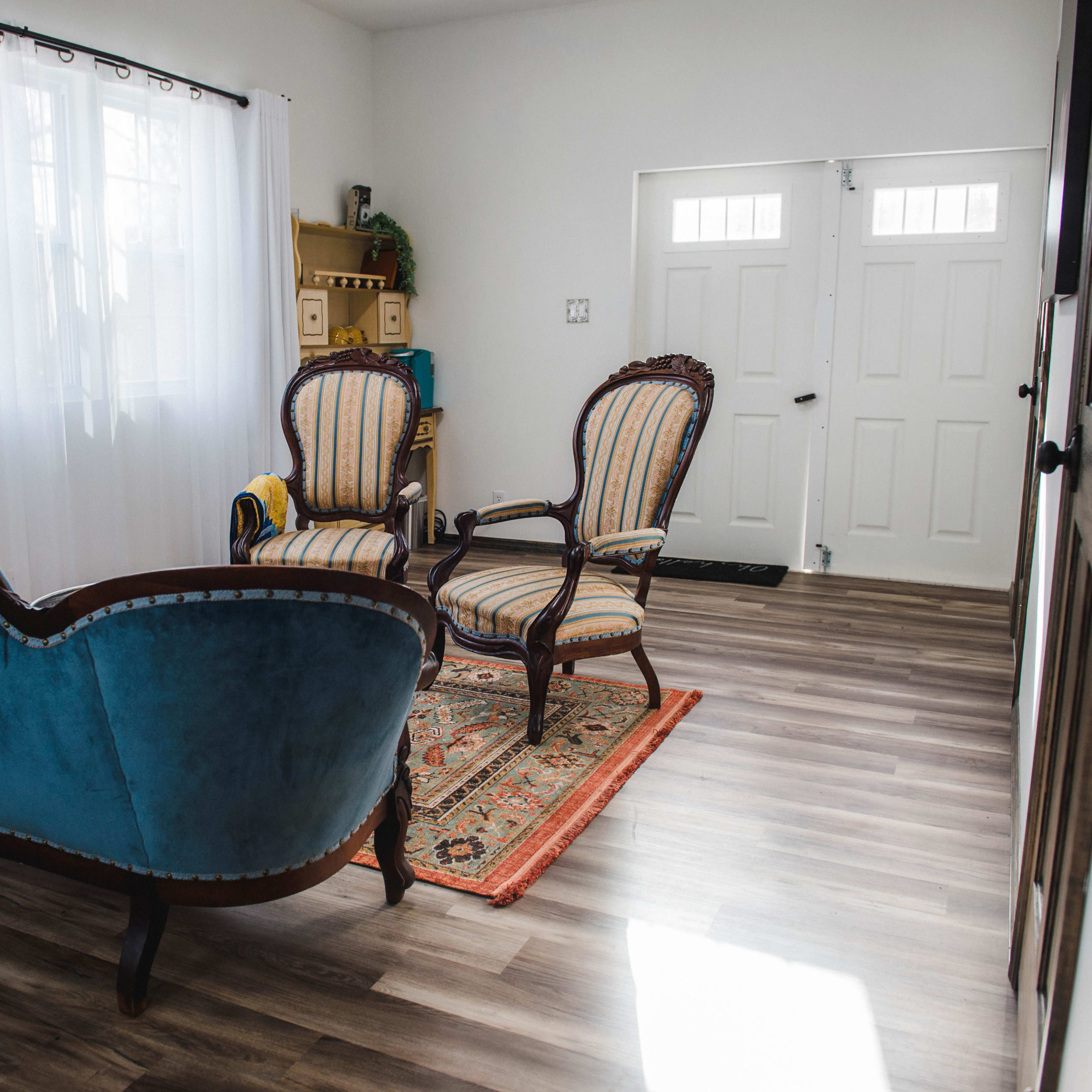 The scene shows a living room with two upholstered chairs and a couch, all arranged atop a patterned rug, leading to a pair of double doors.