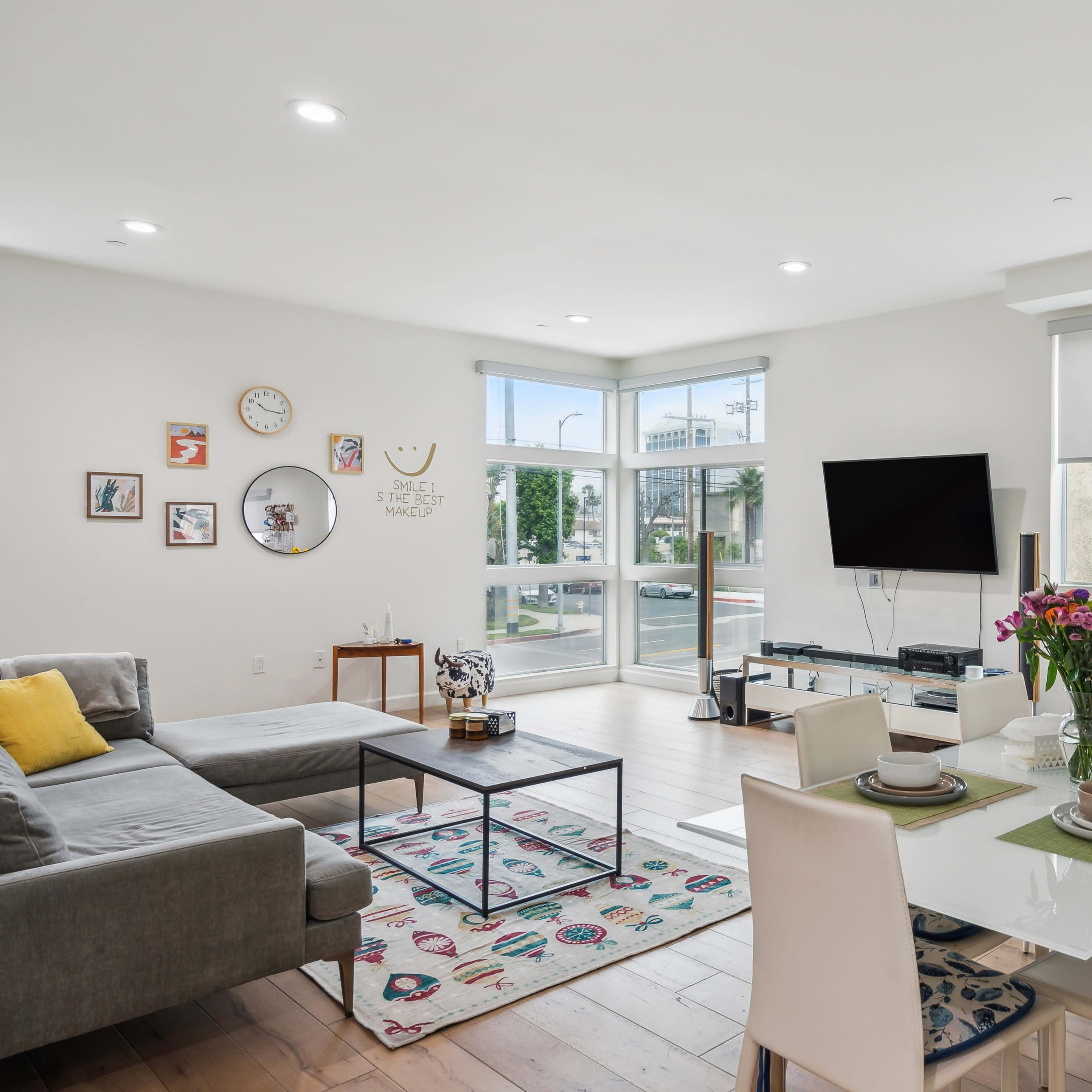 A modern living space features a gray sectional sofa, a dining table set for four, and large windows allowing natural light to fill the room.