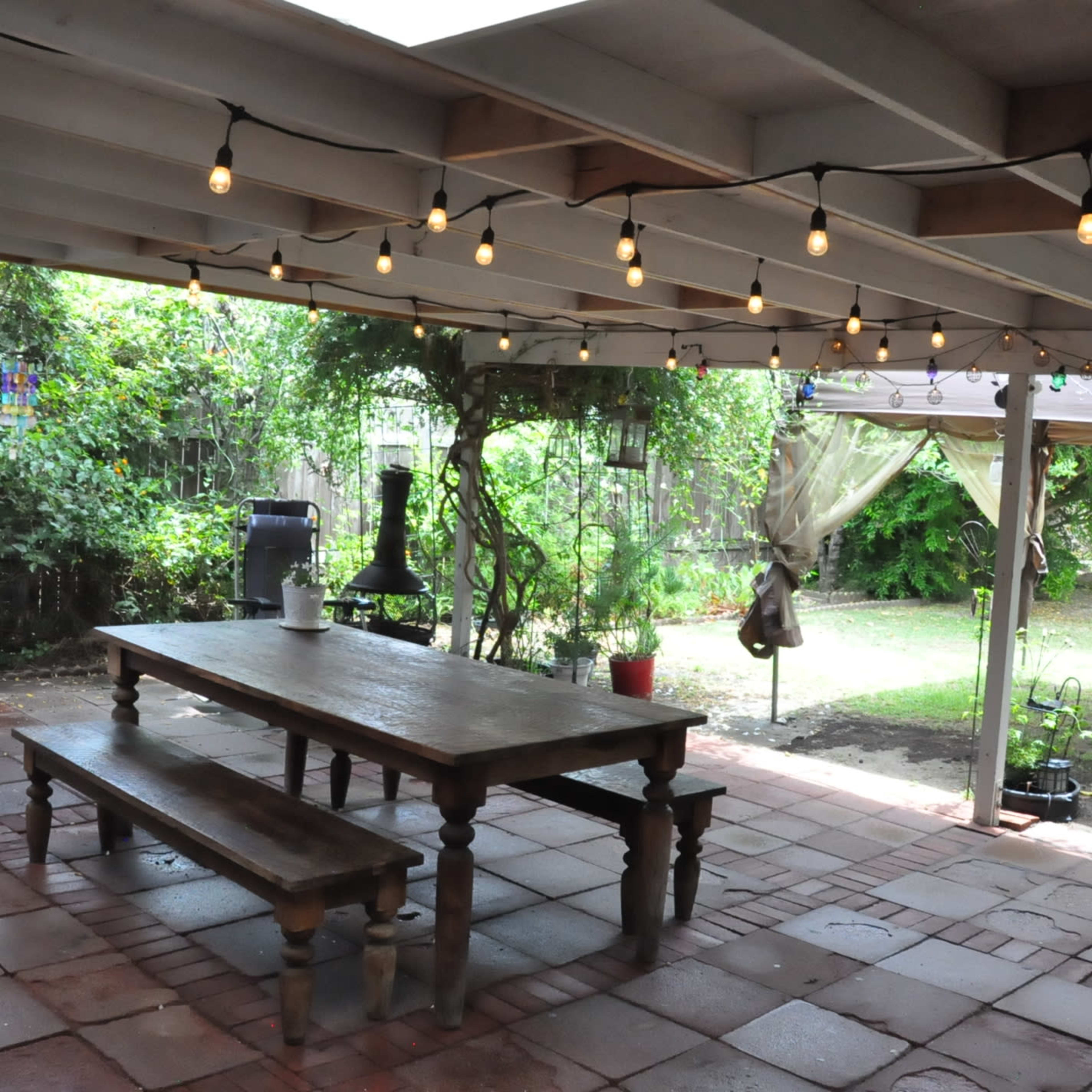 A covered outdoor patio with a long wooden table, string lights overhead, and a garden area in the background.