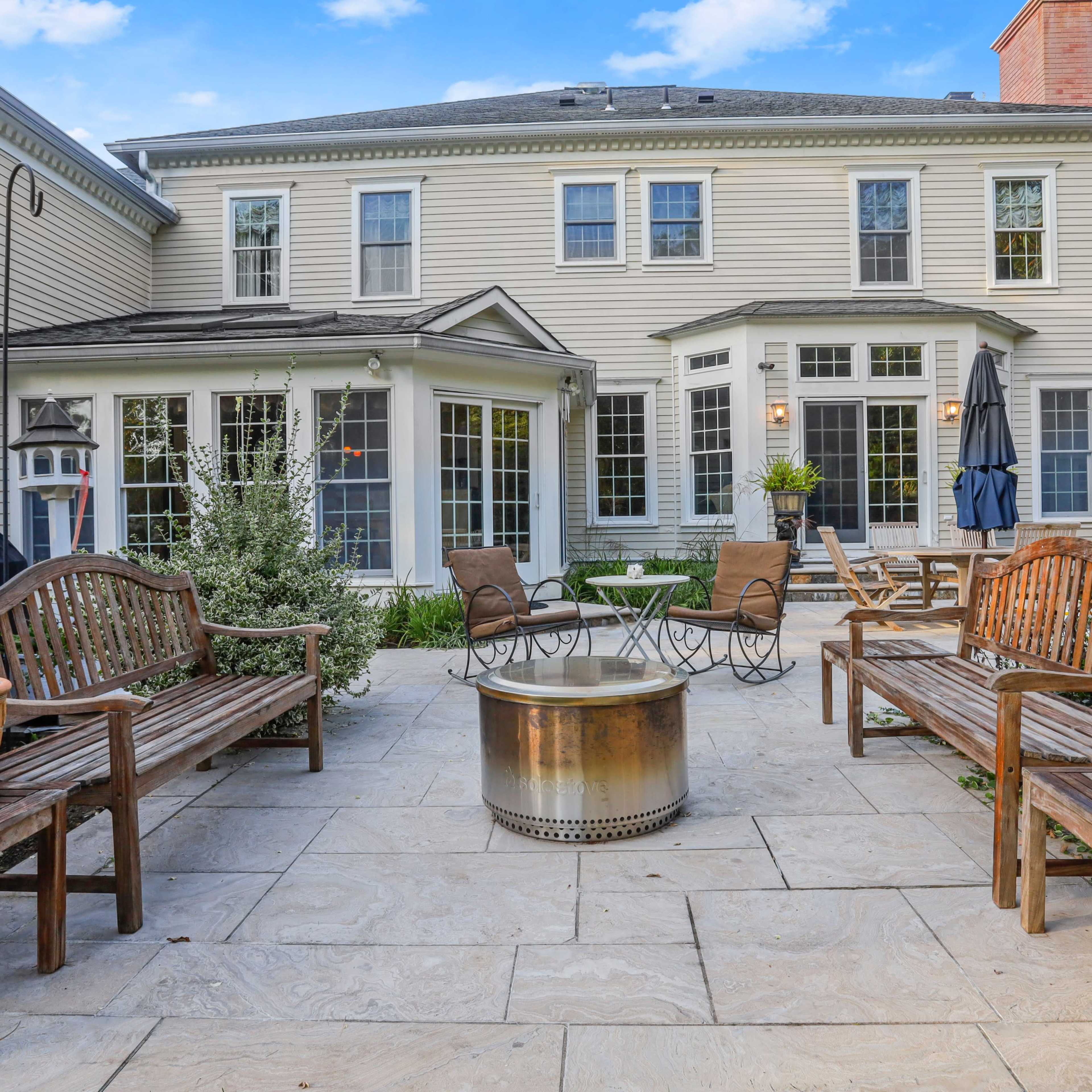 A stone patio area surrounded by wooden benches and a fire pit, with a house featuring multiple windows in the background.