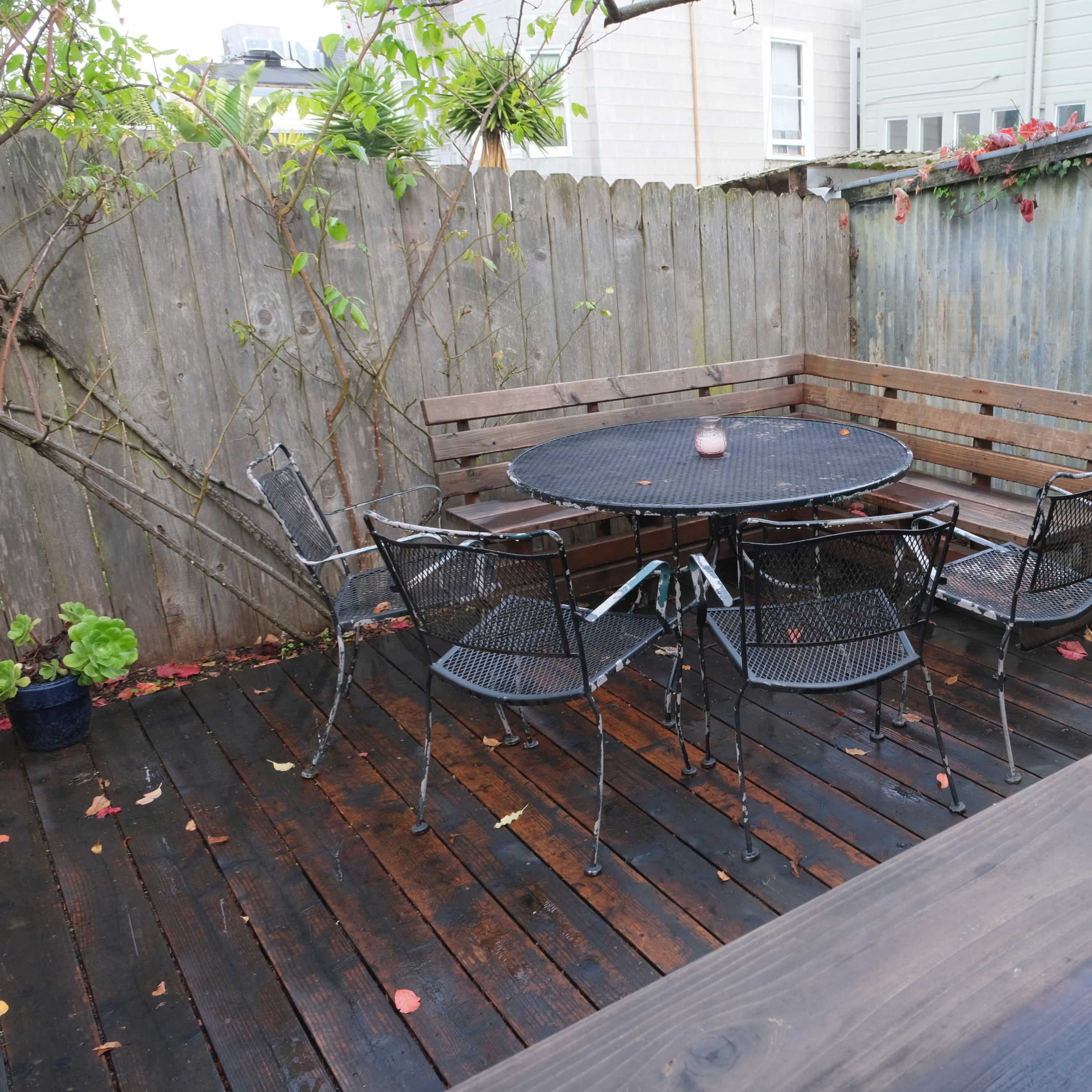 A patio area with a round table surrounded by six metal chairs, set on wooden decking and enclosed by a wooden fence adorned with climbing plants.