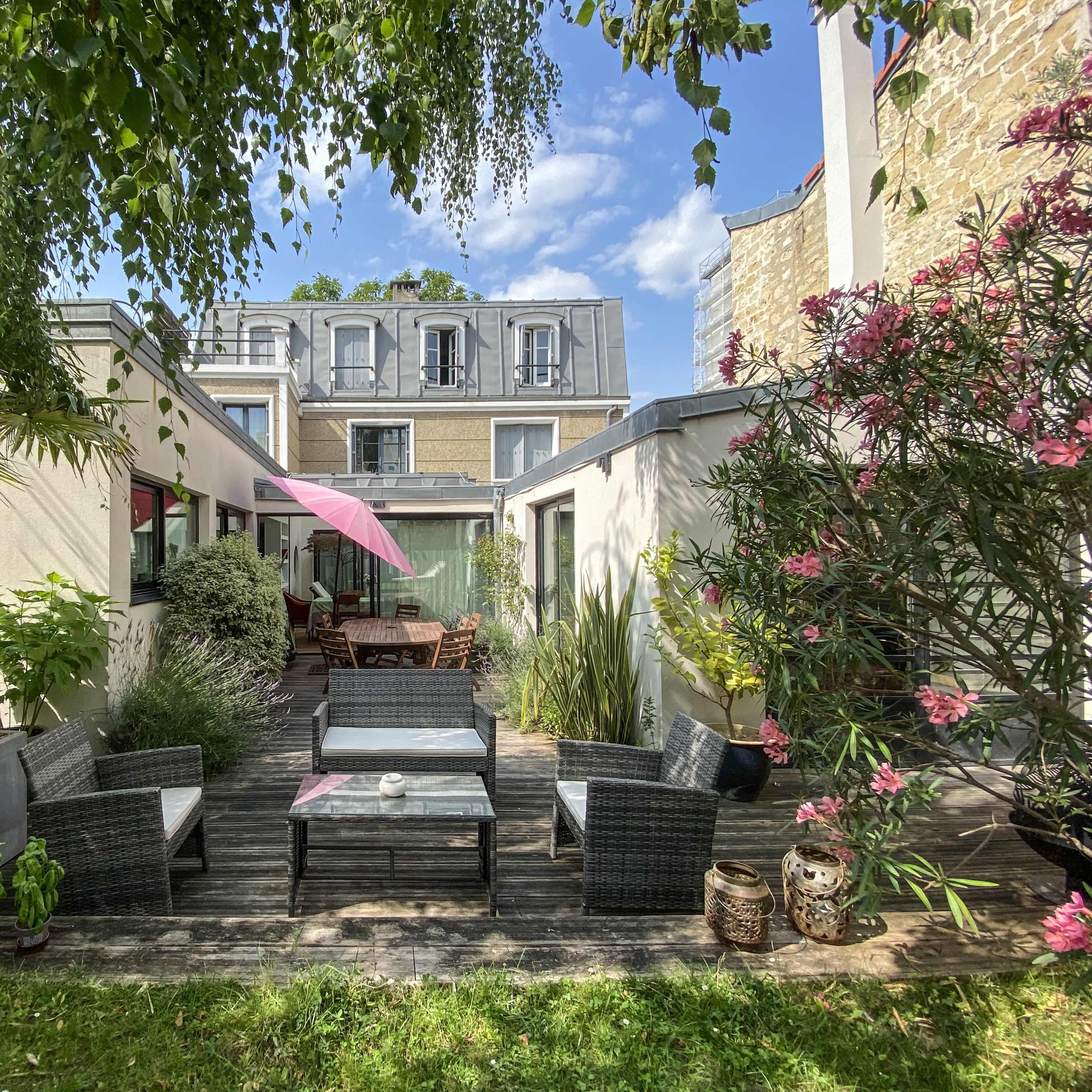A patio area is surrounded by greenery and flowers, featuring outdoor furniture and a pink umbrella, with a building in the background.