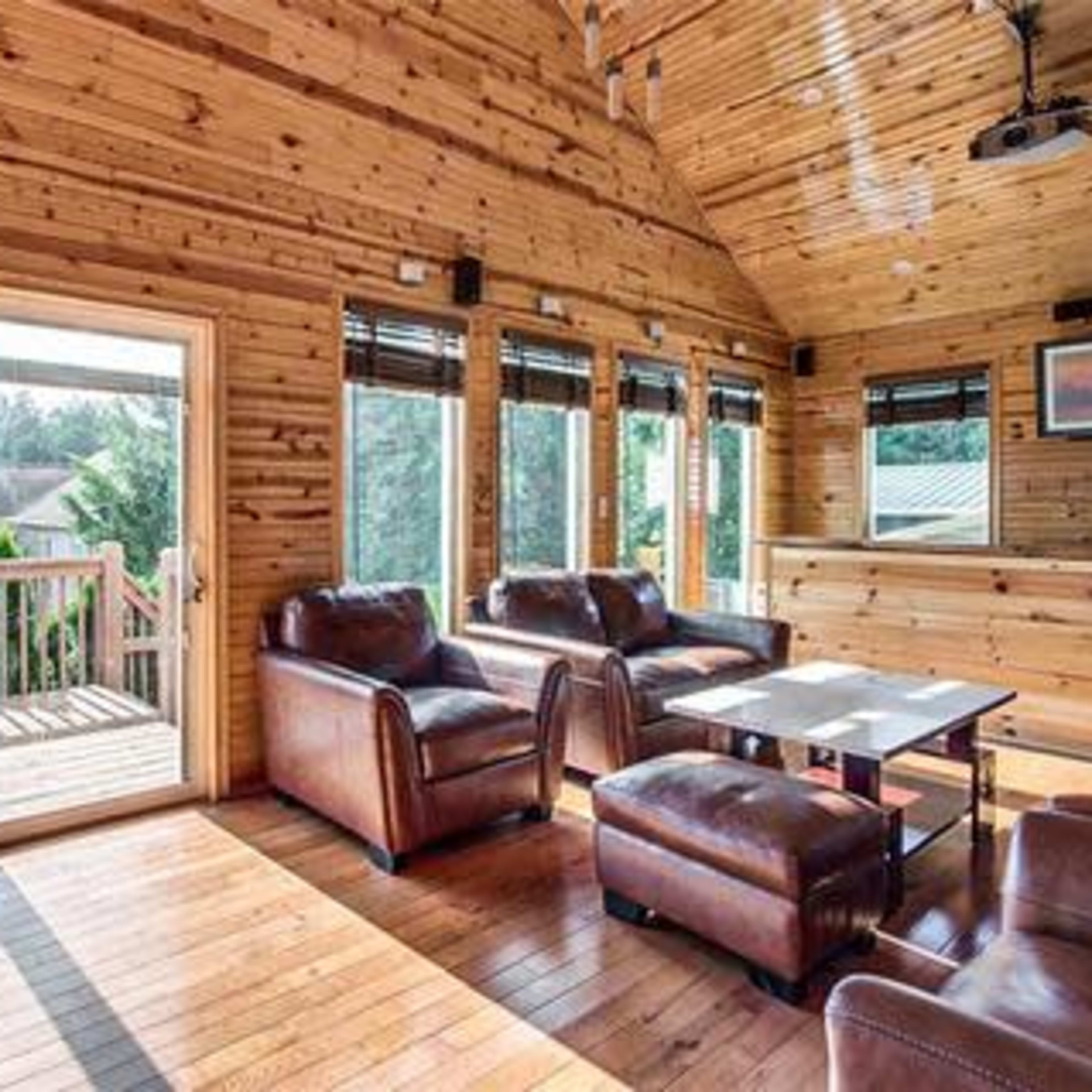 The interior of a wooden-paneled living room features leather couches, a coffee table, and large windows with a view of the outdoors.