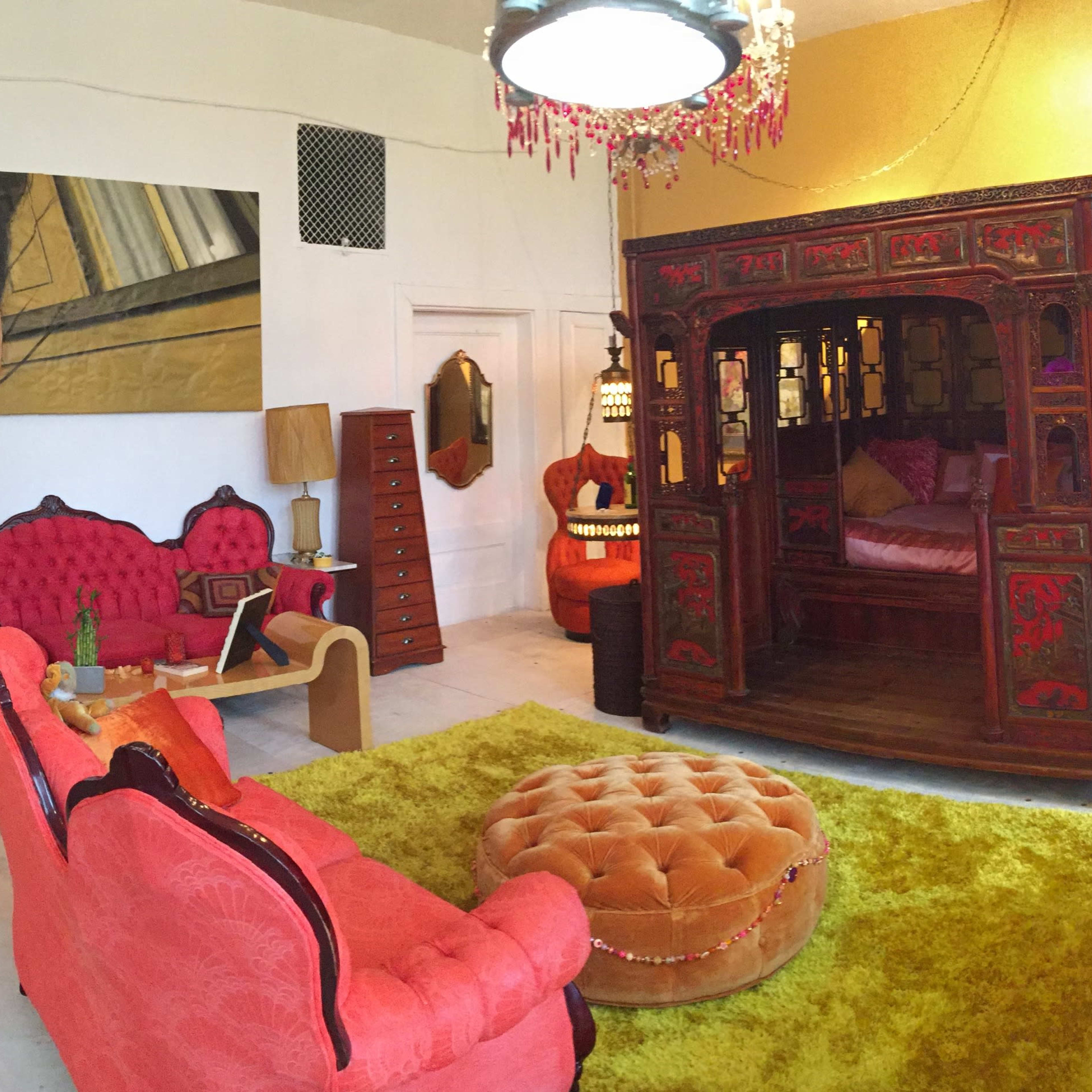 The scene shows a colorful living room featuring ornate red furniture, a vibrant yellow wall, a large wooden bedframe, and various decorative elements.