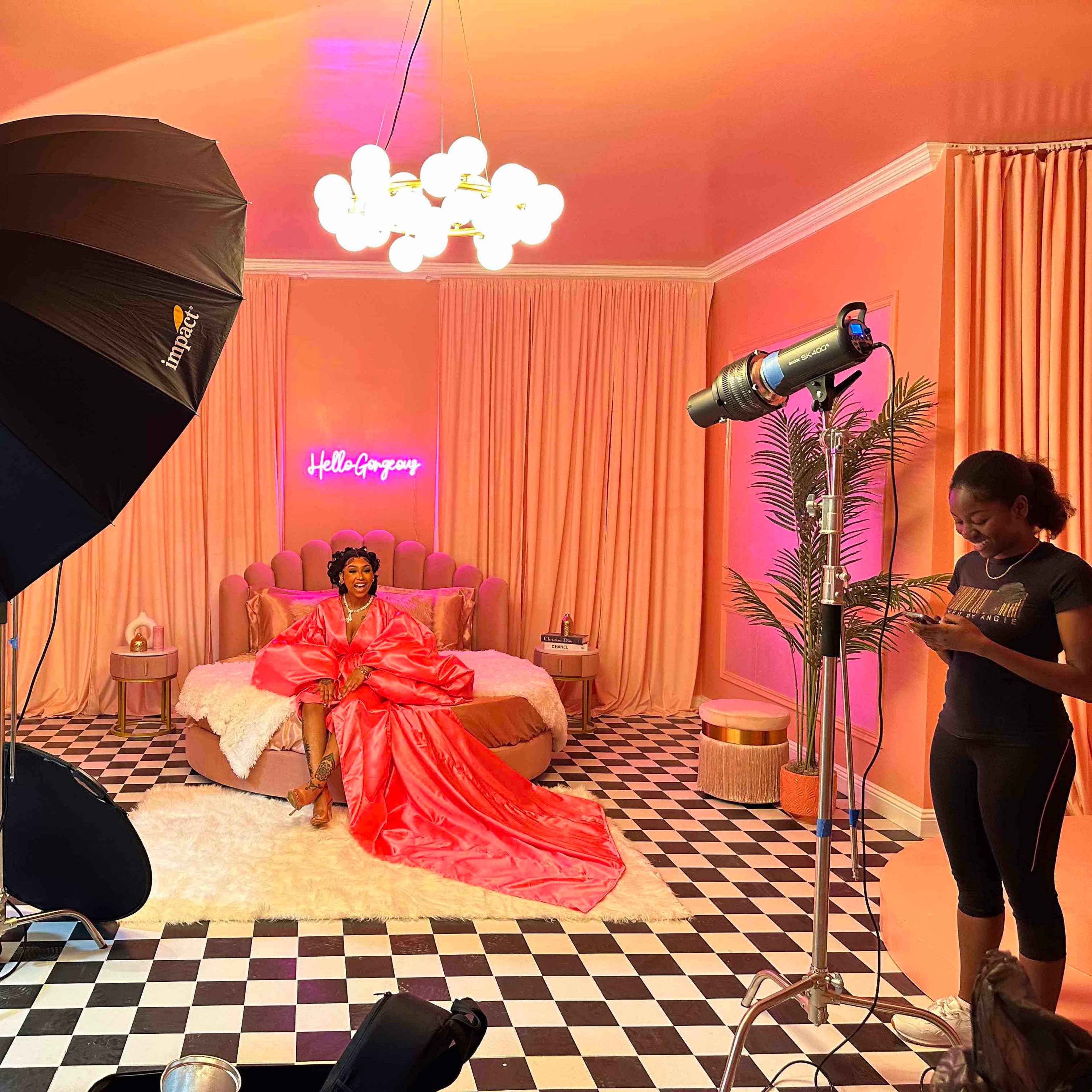 A model dressed in a pink gown poses on a bed in a brightly colored studio with pink walls and decorative lighting.