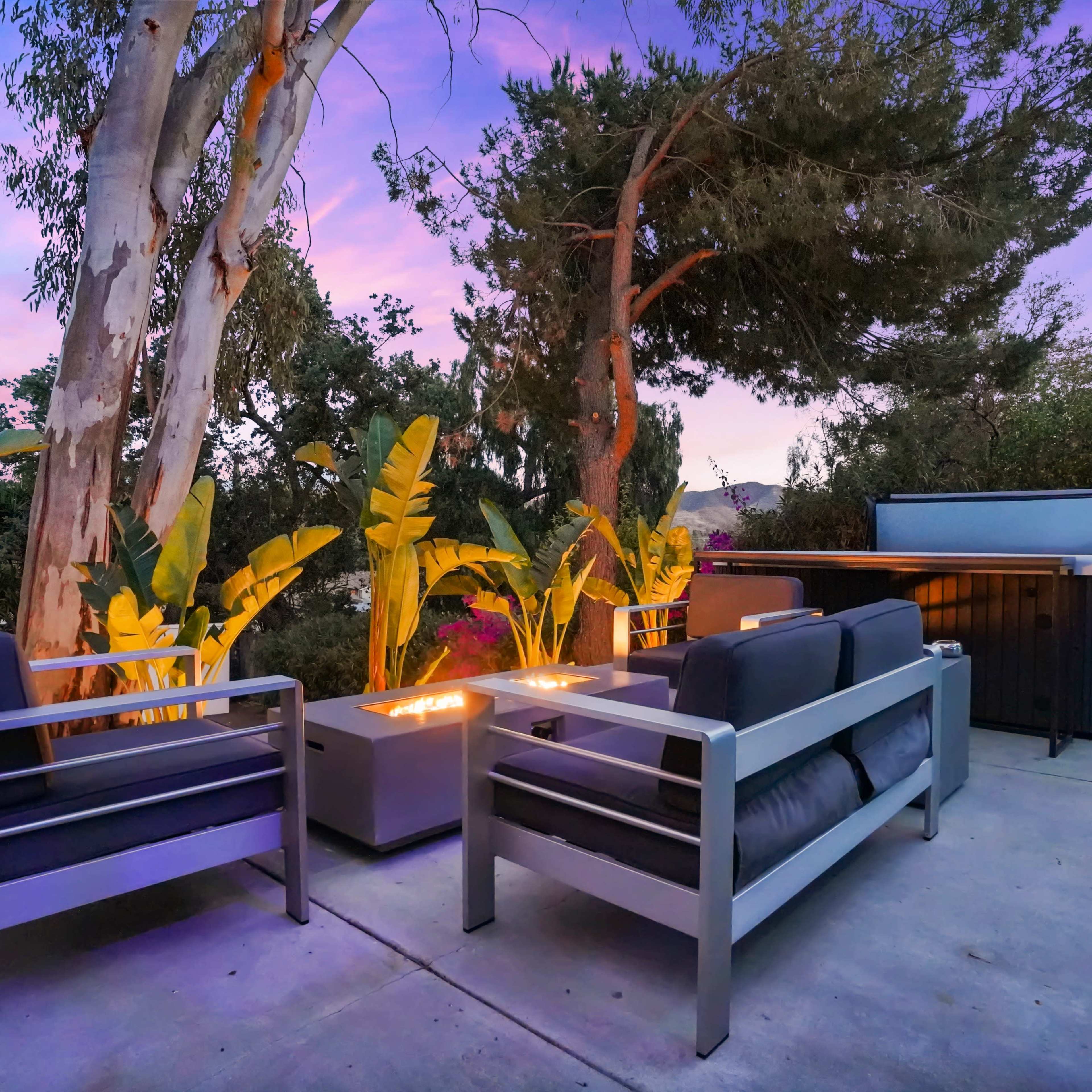 A modern patio features metal-framed seating around a fire pit, surrounded by tropical plants and trees at dusk.