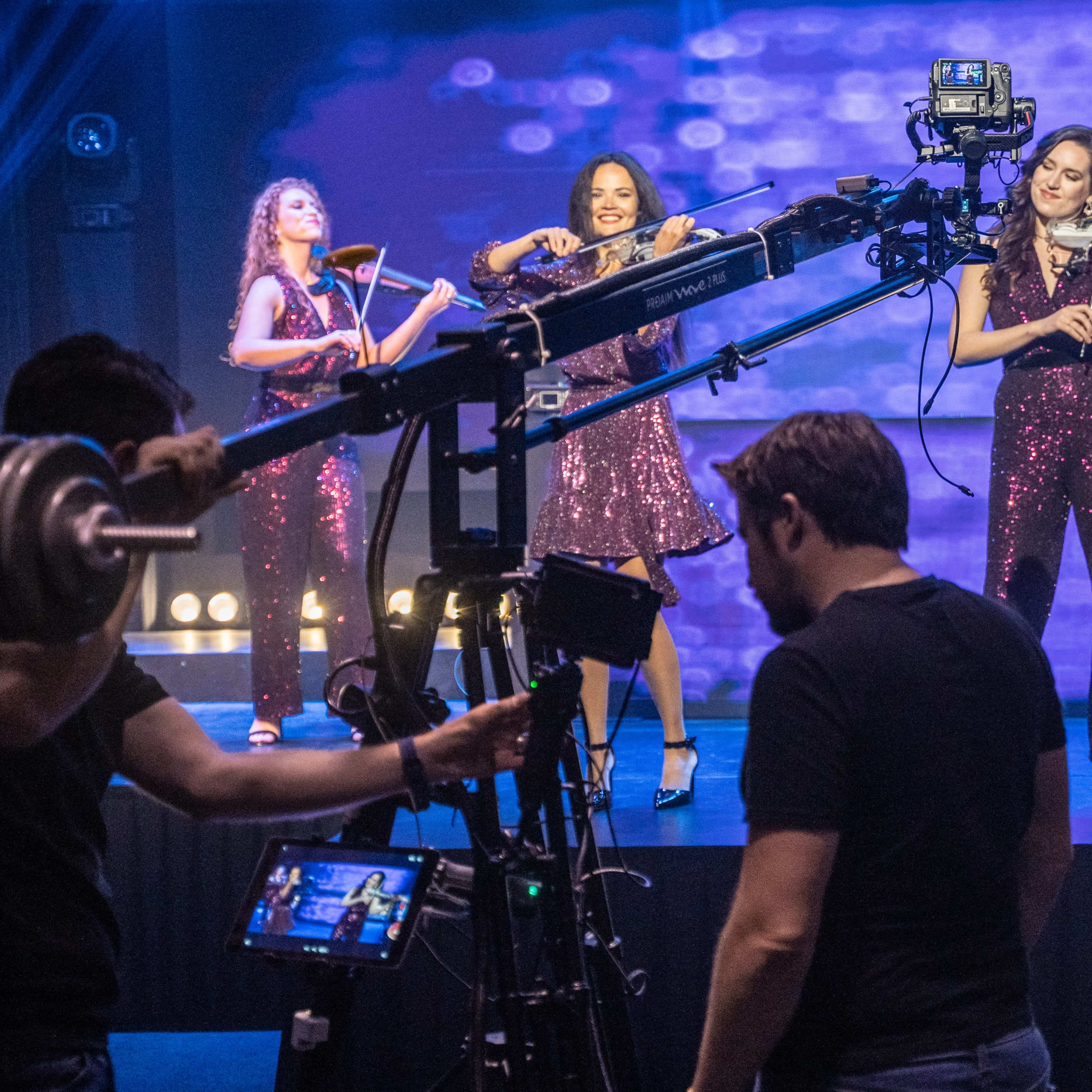 Three women in sequined outfits perform on stage while being filmed by a crew with cameras and lighting equipment.