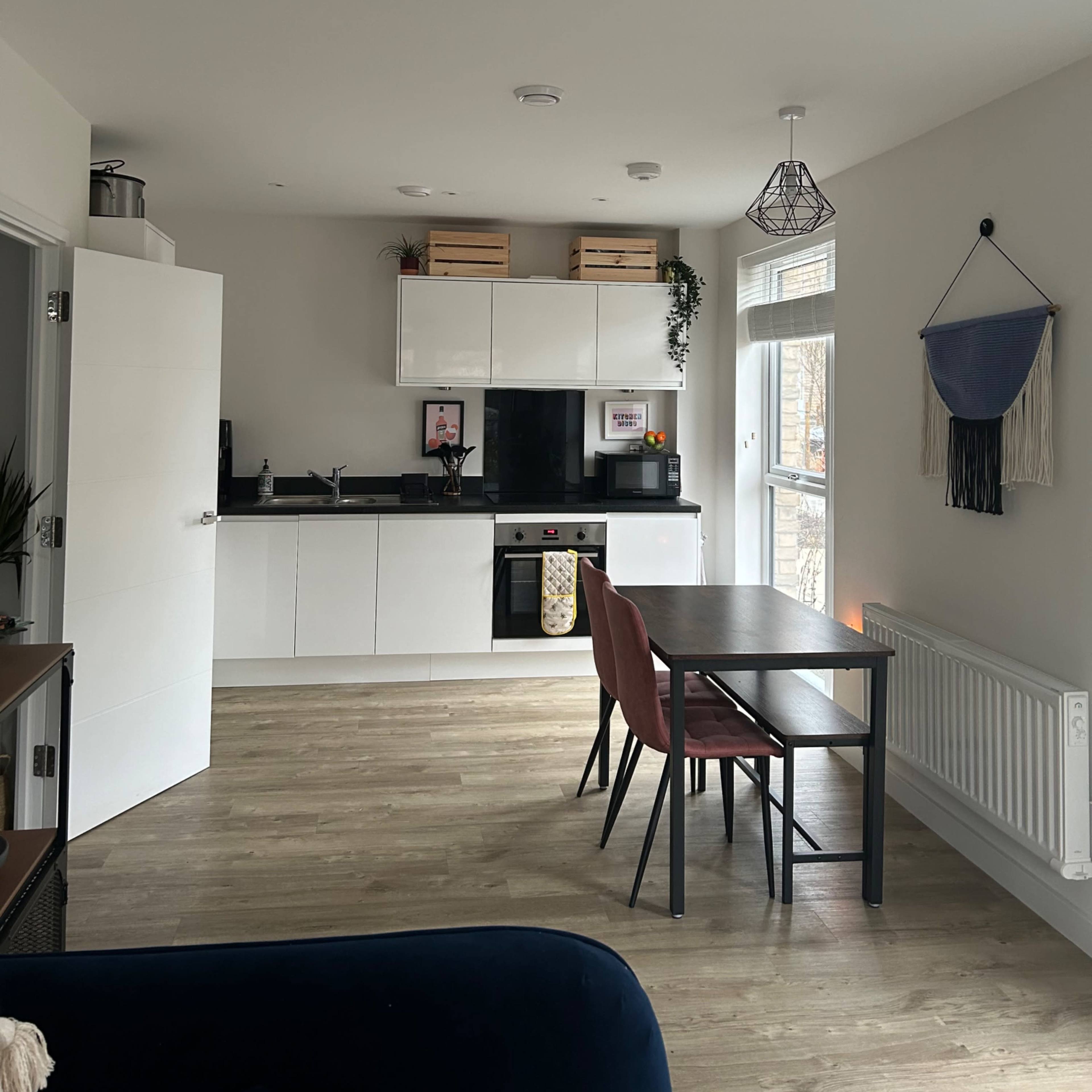 A modern kitchen area features white cabinets, a black countertop, a dining table with chairs, and large windows allowing natural light.