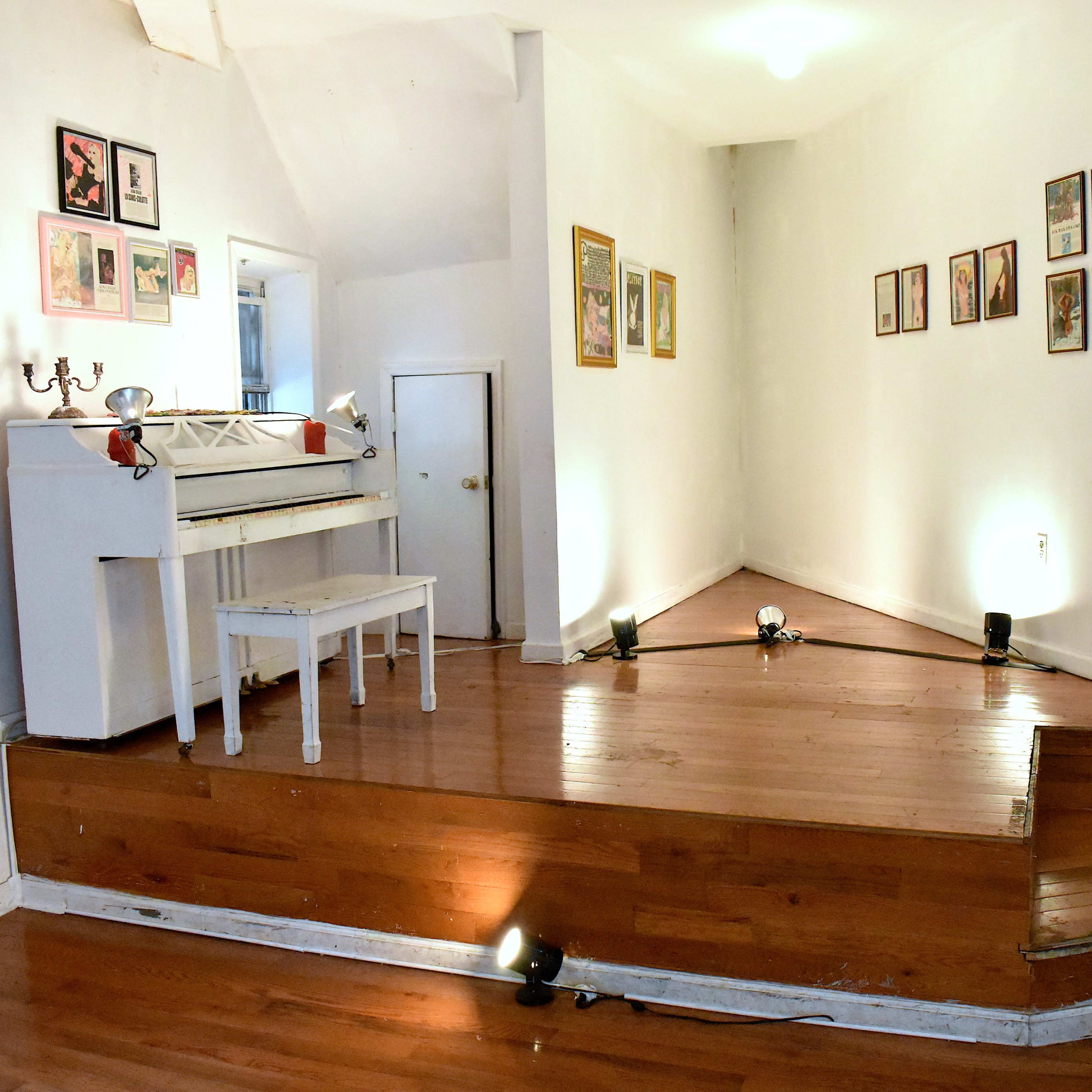 A bright, minimalist room featuring a white piano and a small stool on a wooden platform, with framed pictures arranged on the walls.
