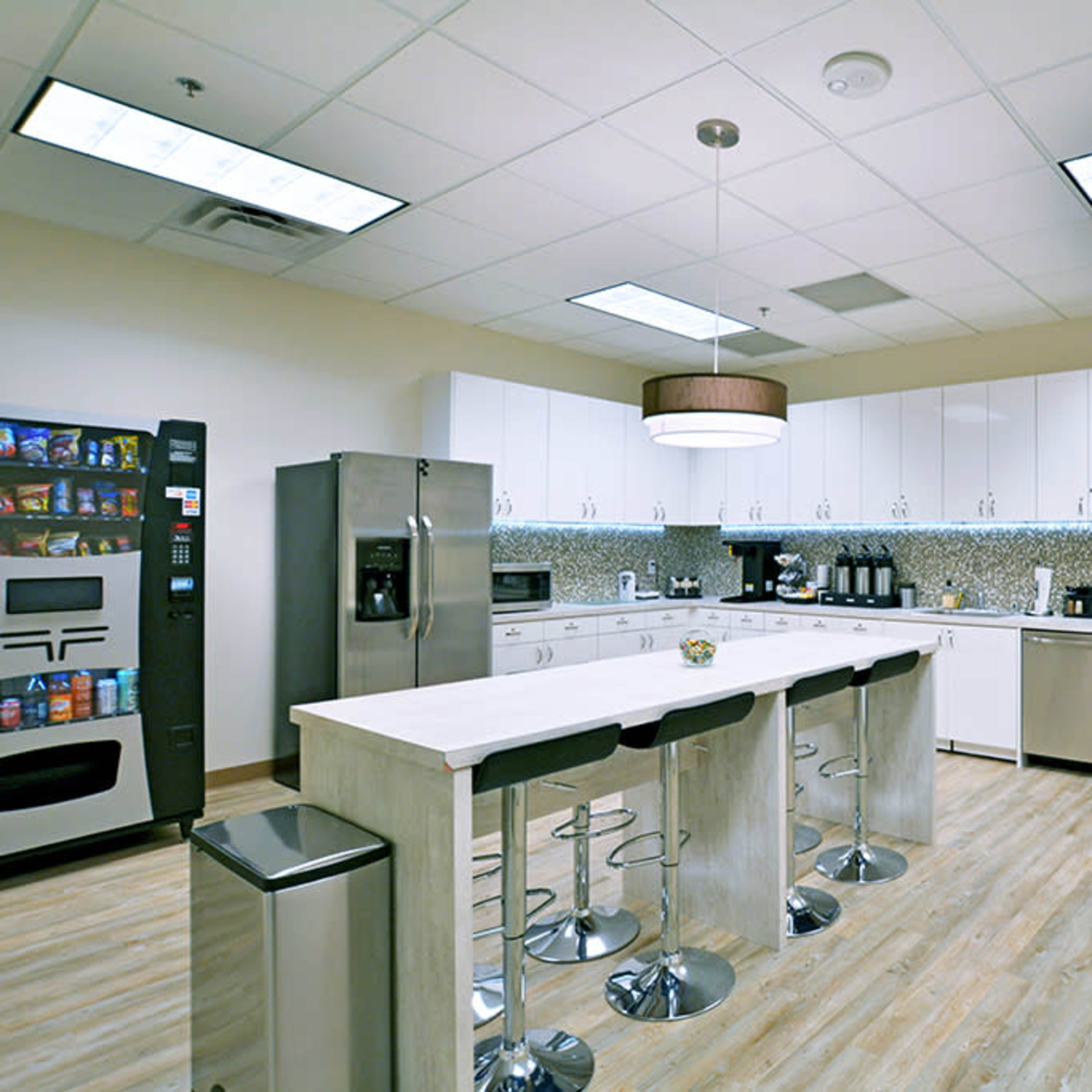 A modern kitchen area equipped with a refrigerator, vending machine, coffee station, and an island with bar stools.