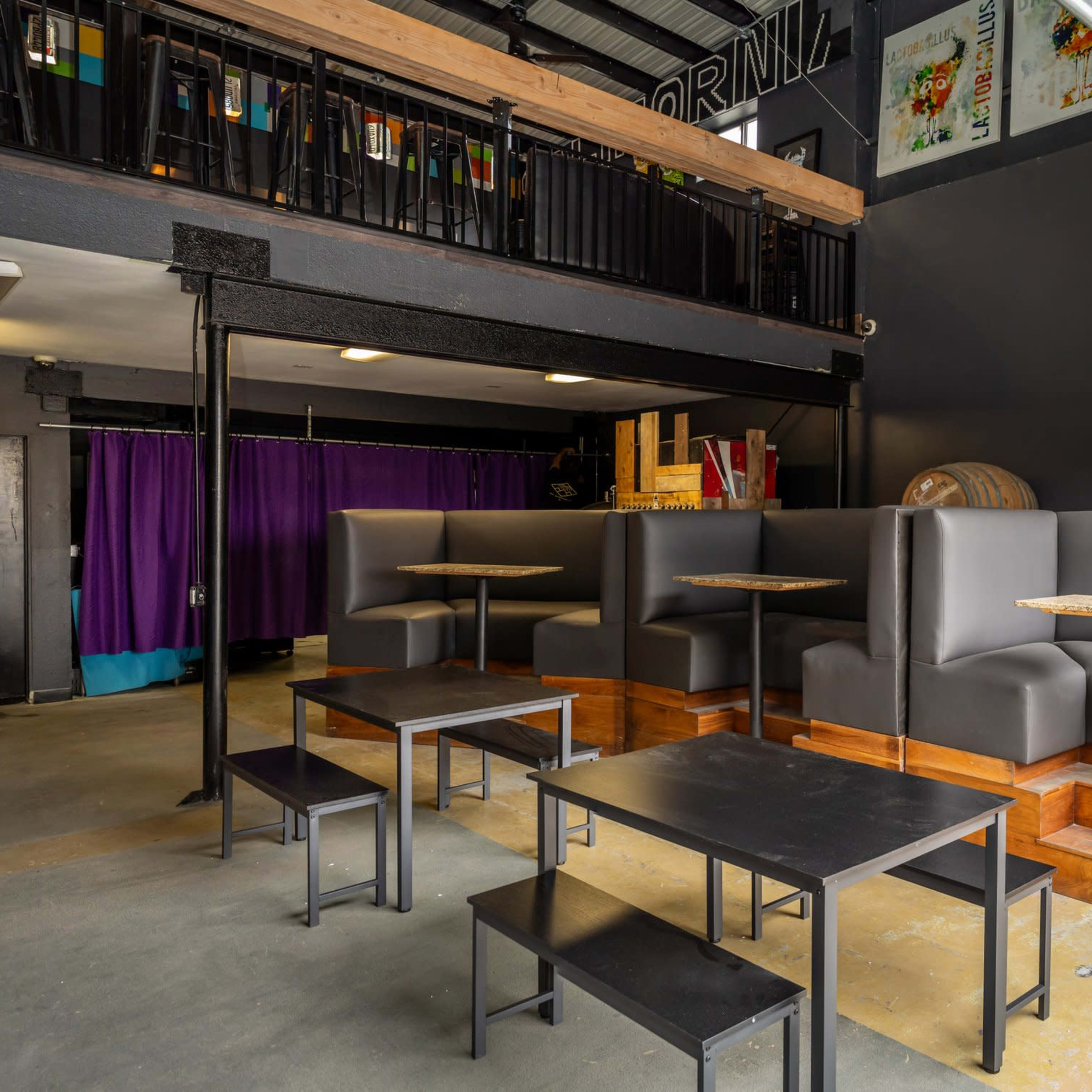 A modern café interior with dark tables and booths, a purple curtain in the background, and a mezzanine with colorful chairs.