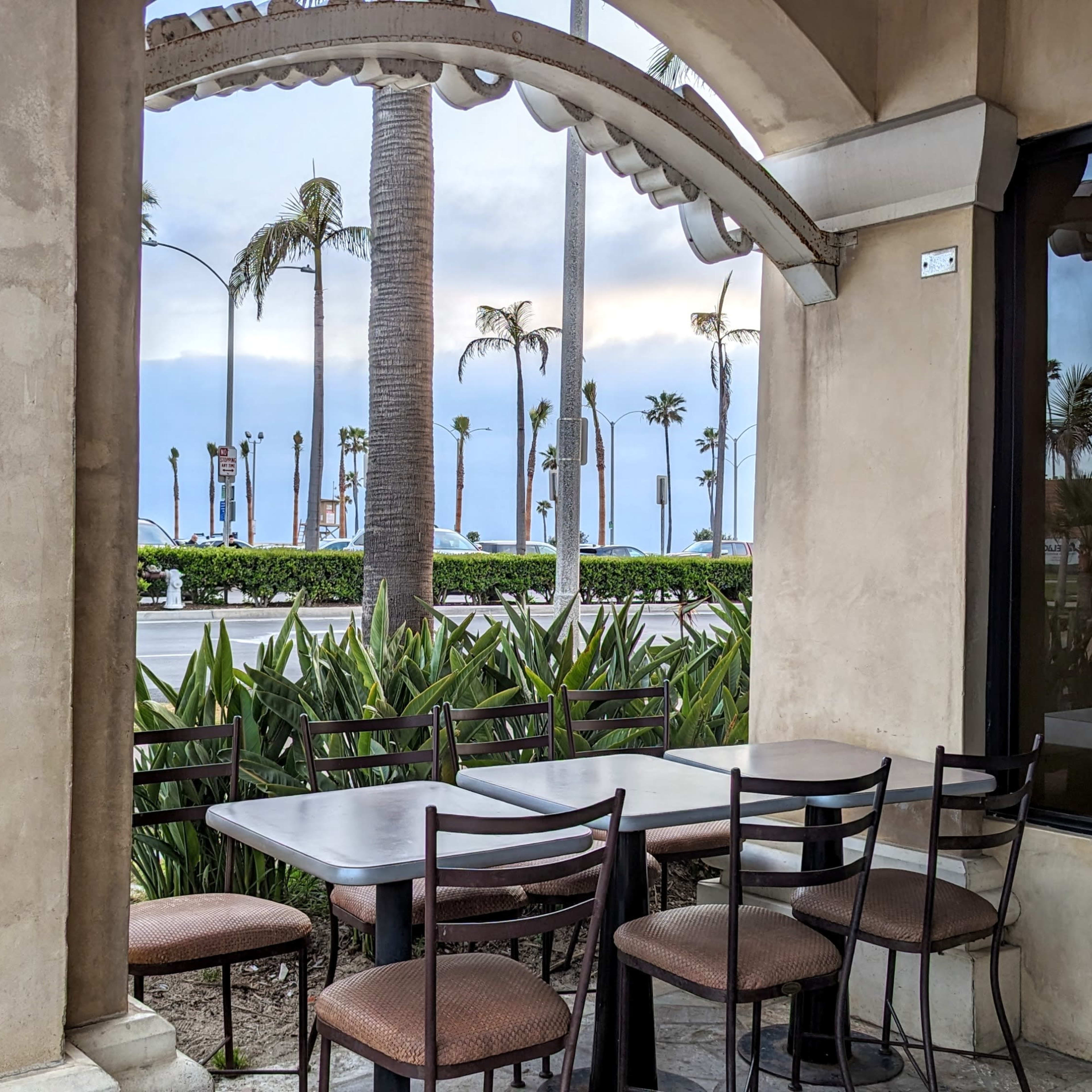 An outdoor seating area with several metal-framed chairs and a table overlooks a palm-lined street.