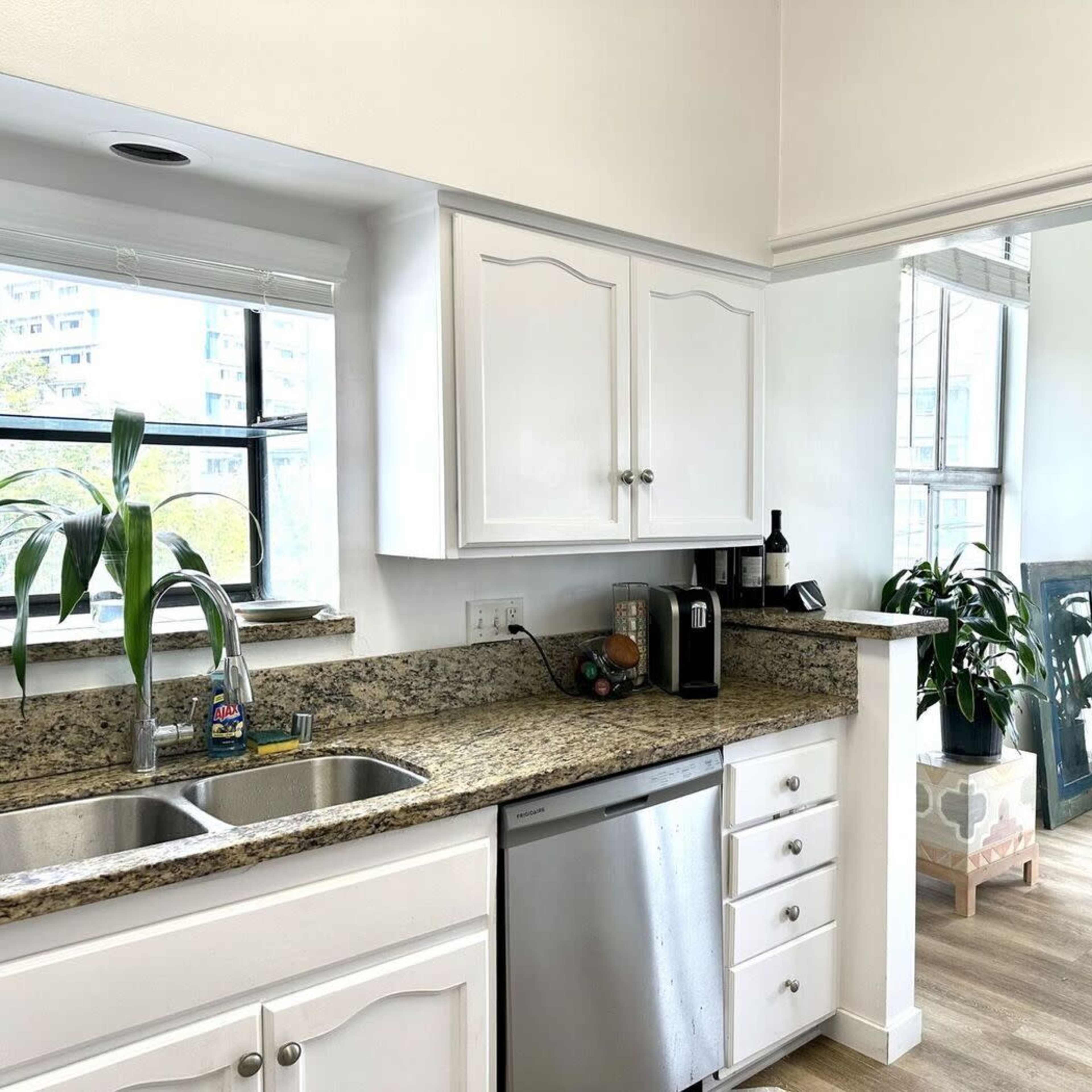 A modern kitchen with white cabinets, stainless steel appliances, and a view of a bright room with plants through a large window.