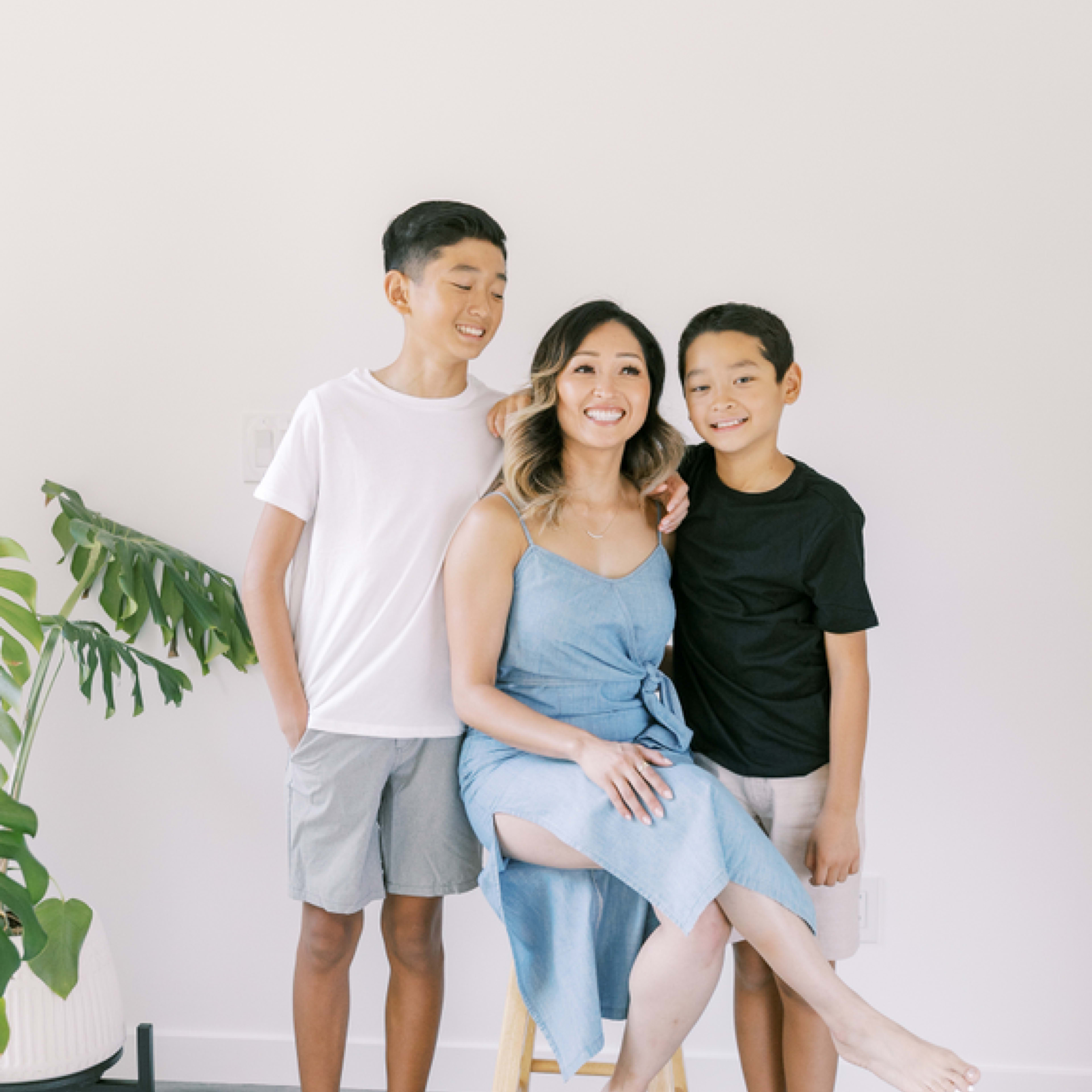 A woman sits on a stool with two boys standing beside her in a bright, minimalist room.
