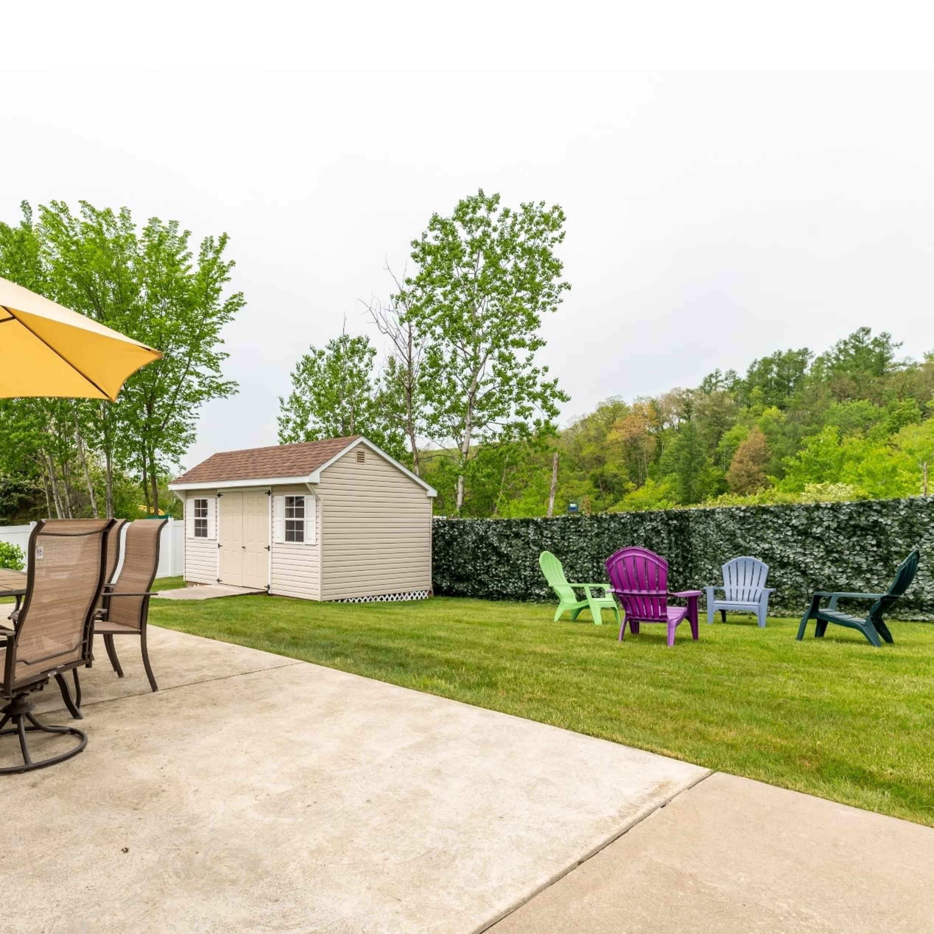 A concrete patio features outdoor furniture and an umbrella, with a shed and colorful chairs set on a grassy yard surrounded by trees.