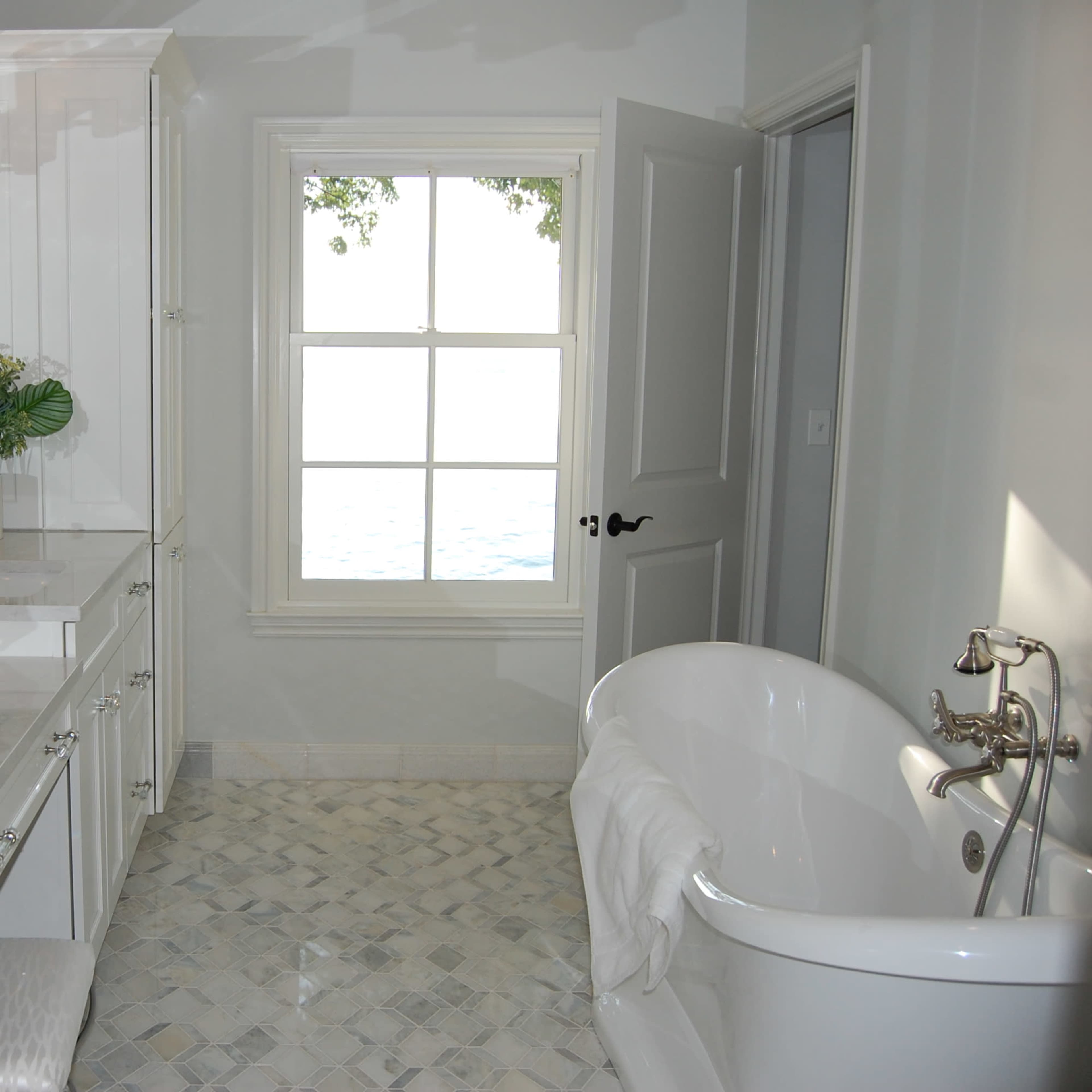 The bathroom features a white freestanding tub, double sinks with marble countertops, and a window overlooking a body of water.