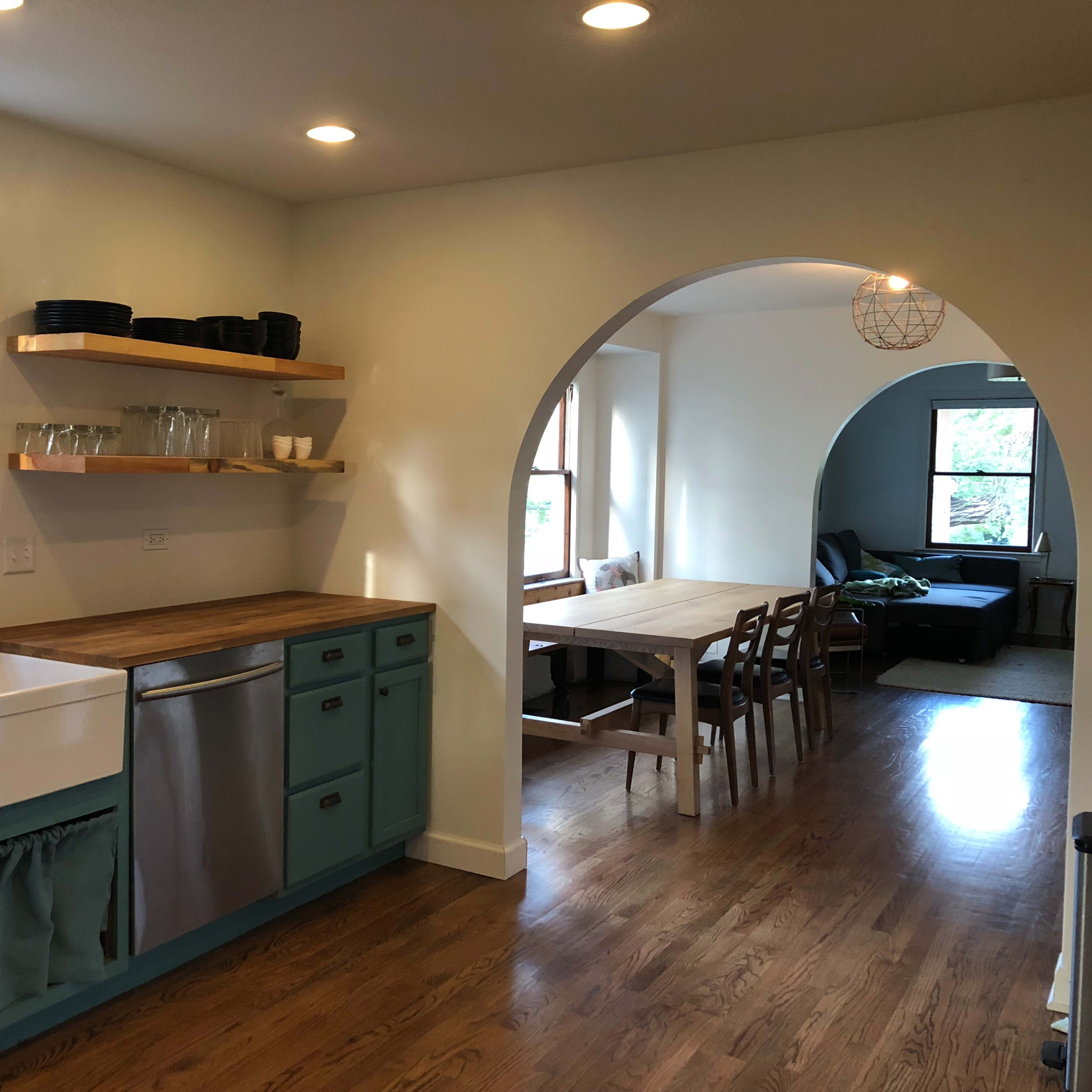 A kitchen with a farmhouse sink and wooden countertops opens to a dining area with a table and chairs, framed by a large archway.