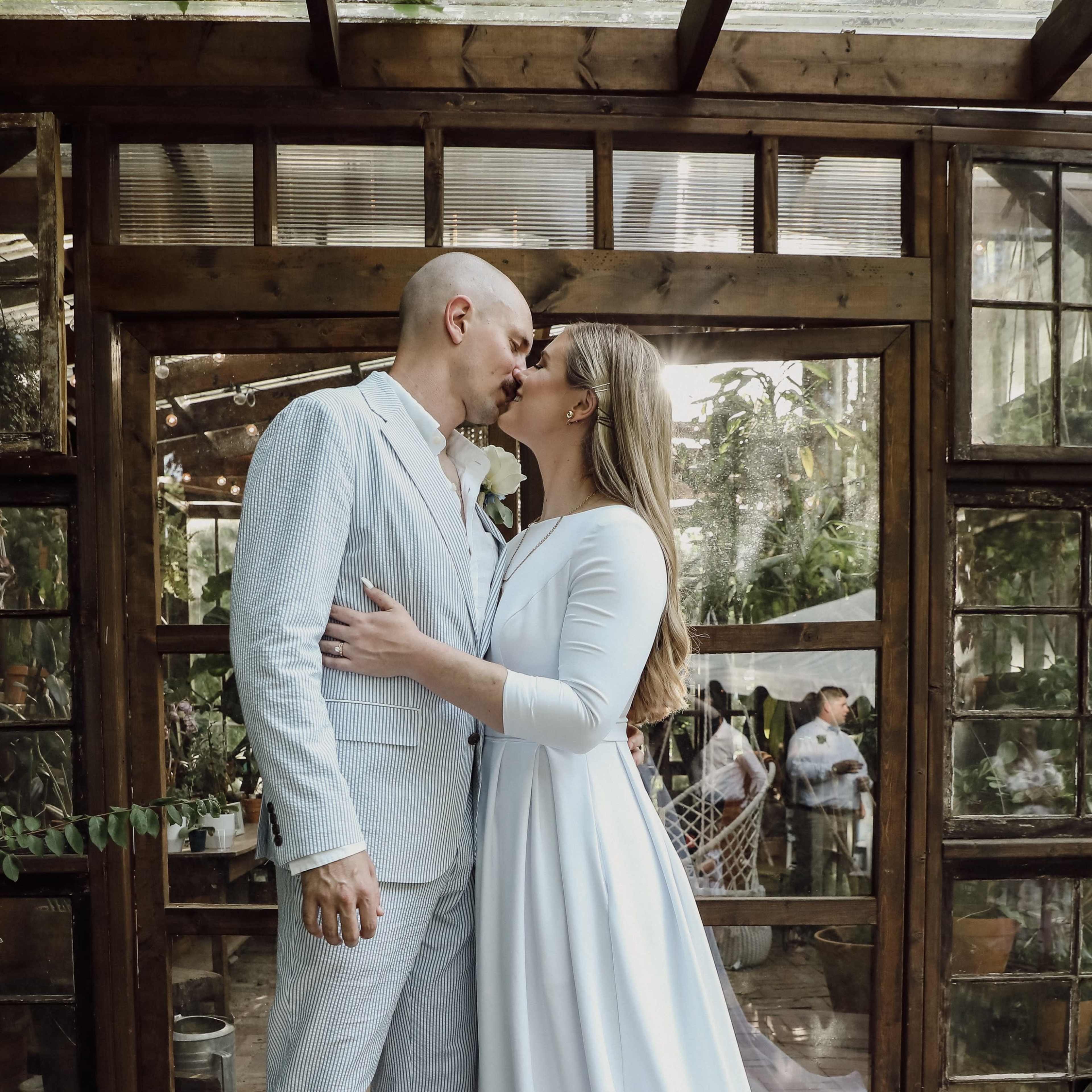 A couple shares a kiss in an indoor garden setting, surrounded by glass walls and greenery.