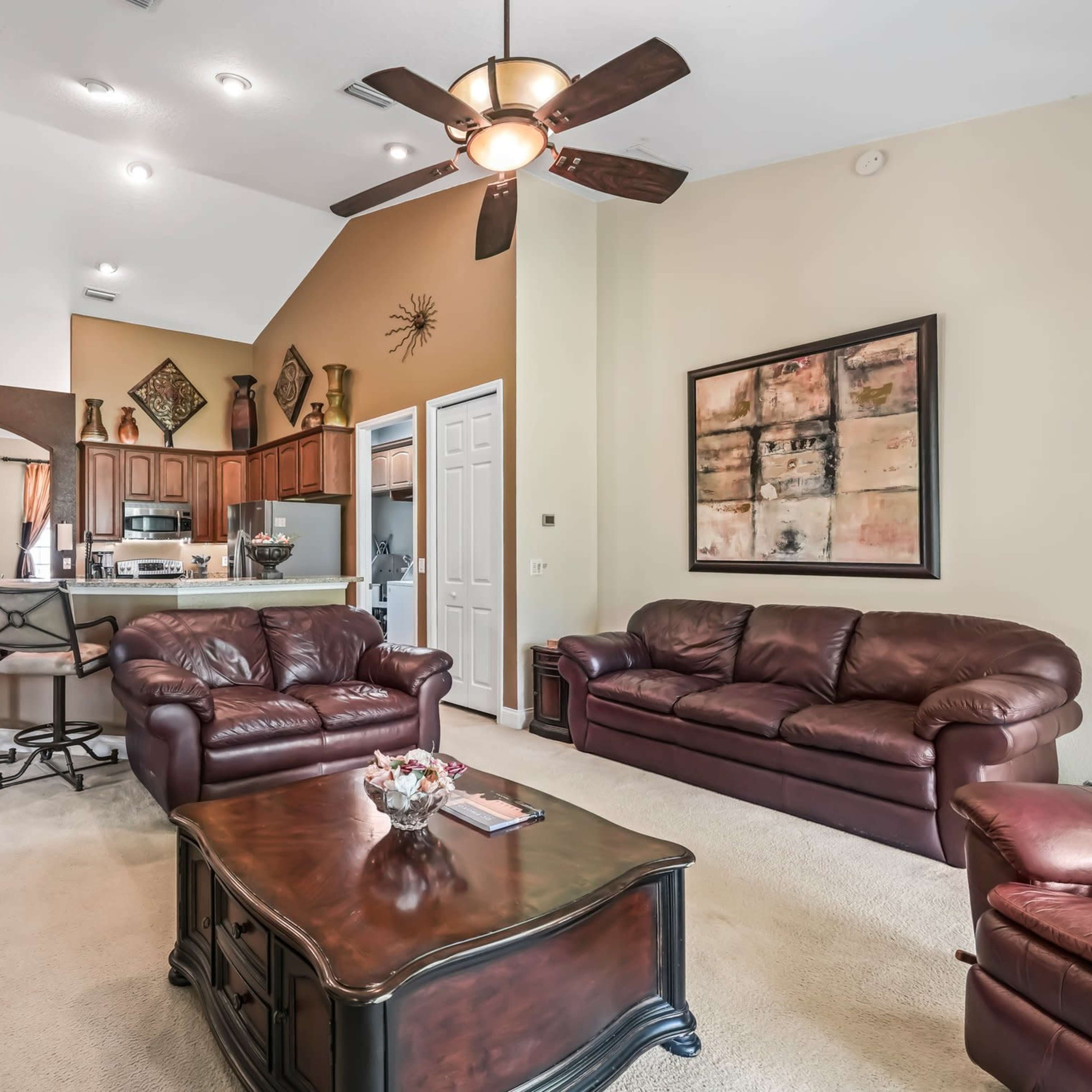 A spacious living room featuring two brown leather couches, a coffee table, and an open kitchen area in the background with wooden cabinets.